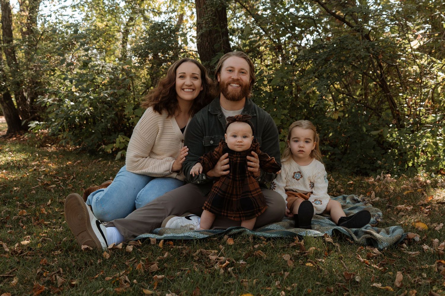Family of four posing together on a blanket in a lush green autumn park, smiling warmly. Family photography in Regina Saskatchewan.