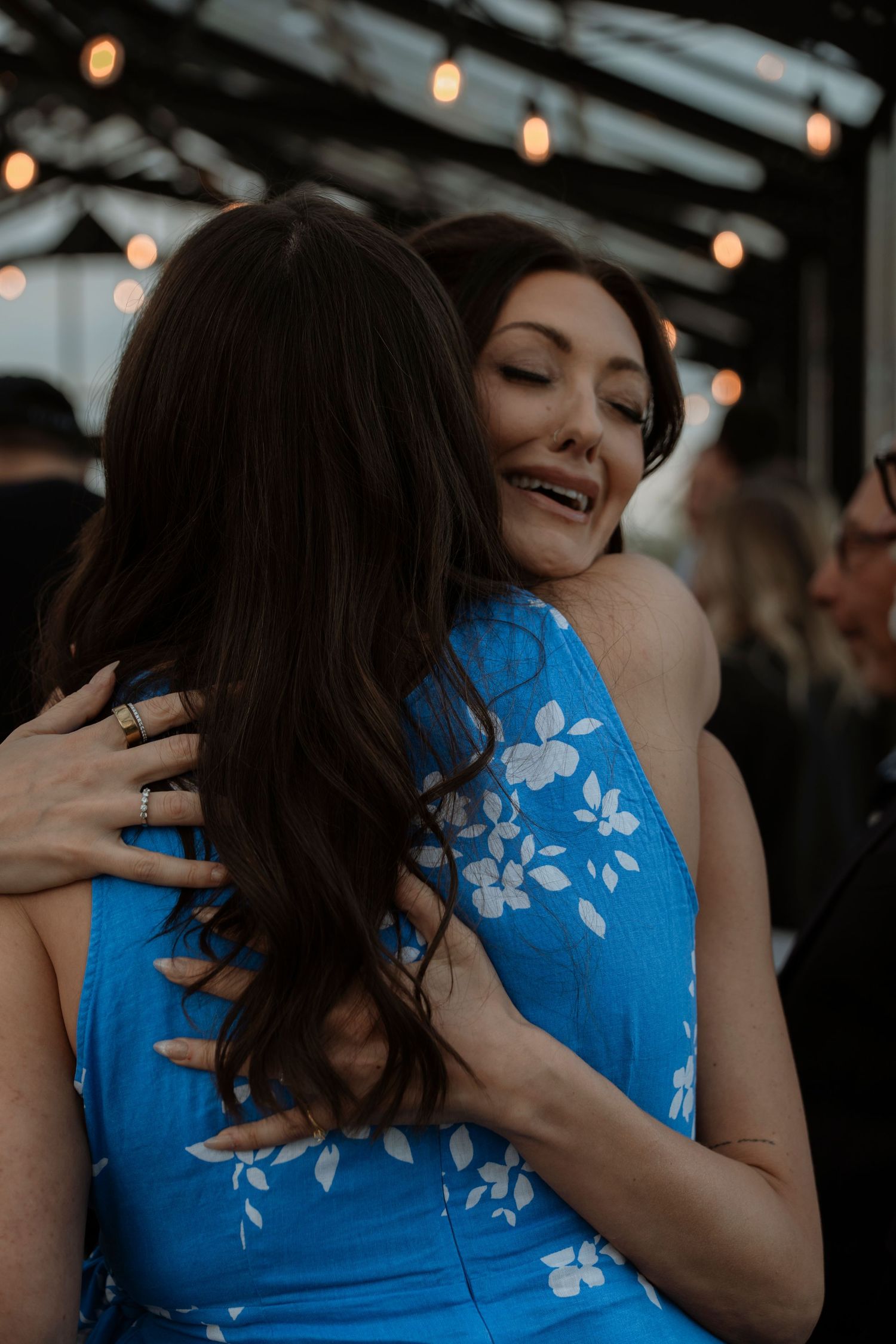 Two women sharing a warm hug at an event, one wearing a blue floral dress, with string lights in the background.