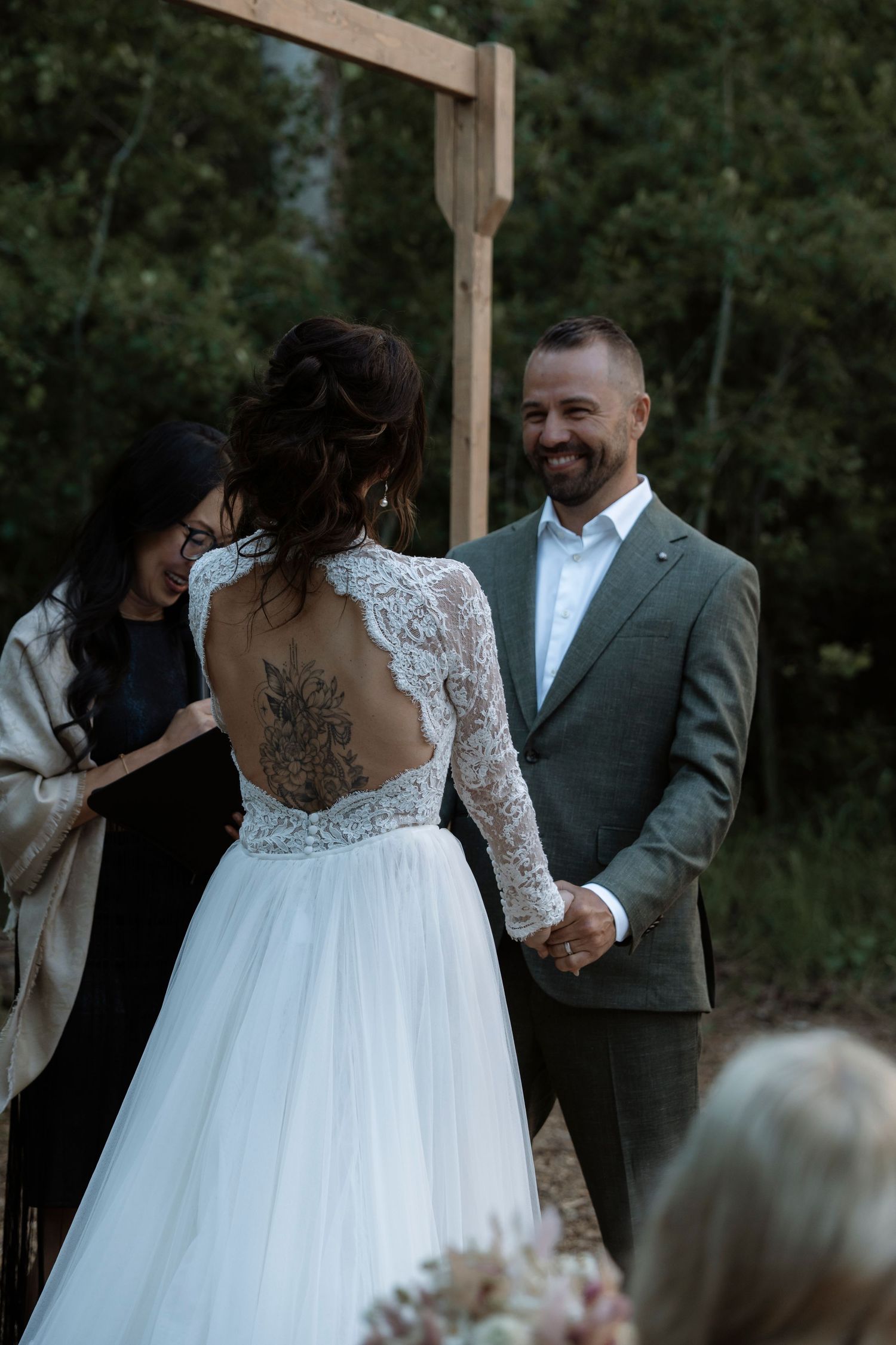 Bride in lace backless wedding dress with floral tattoo holds hands with smiling groom in gray suit at outdoor ceremony.