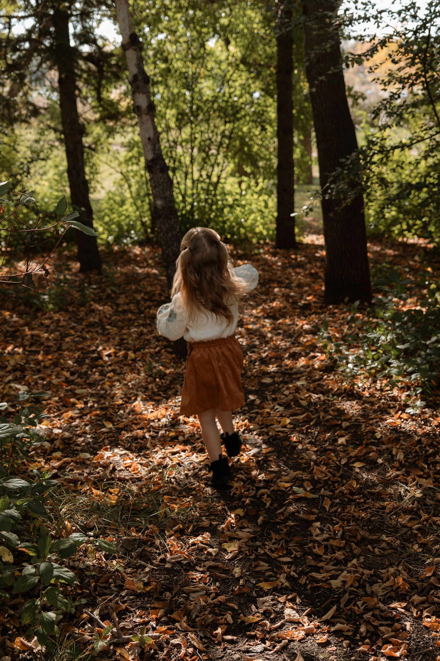 A young girl in a white top and brown skirt runs through a sun-dappled forest covered in autumn leaves. Family photography in Regina Sask.