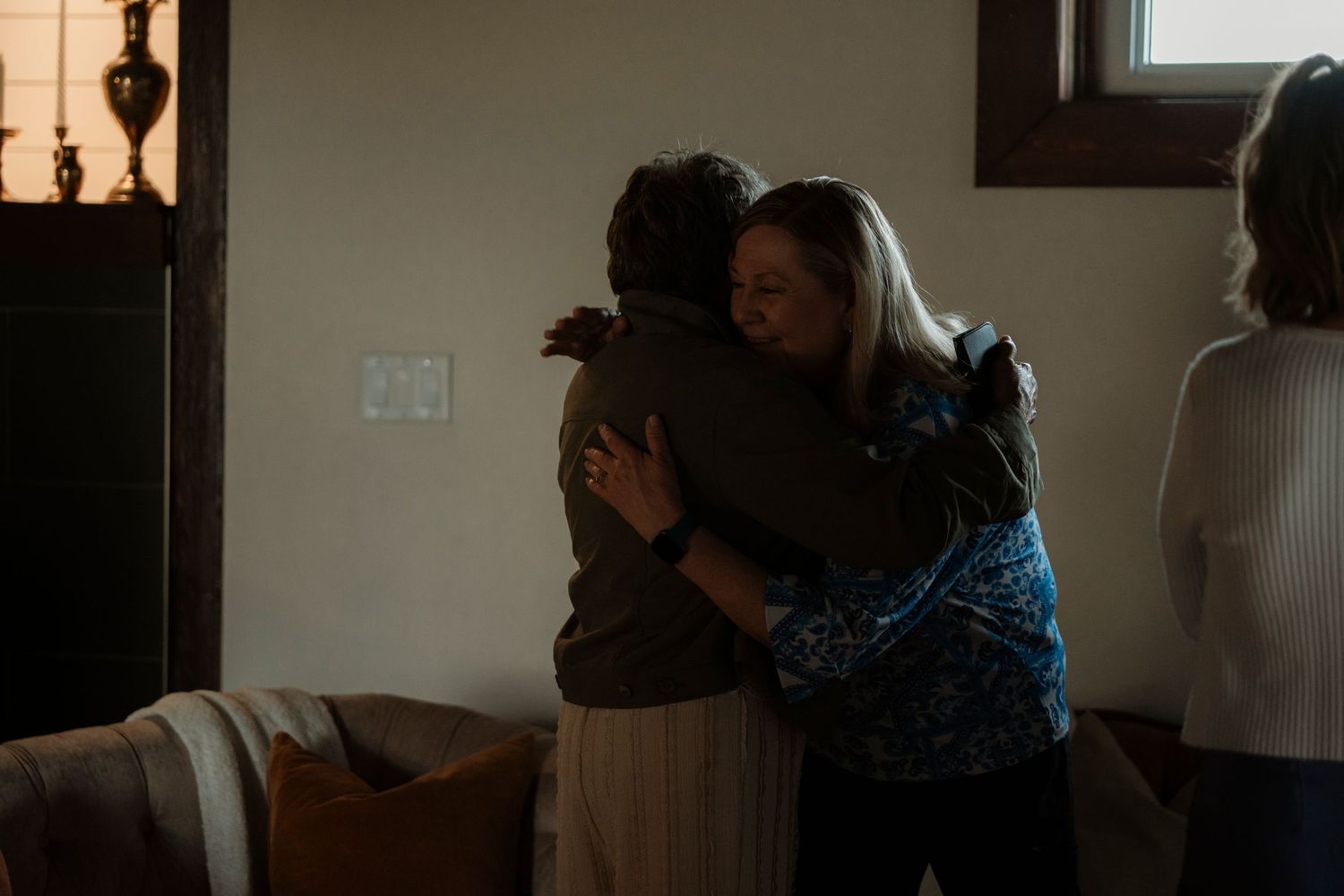 Two people share a warm embrace in a dimly lit living room while another person stands nearby.