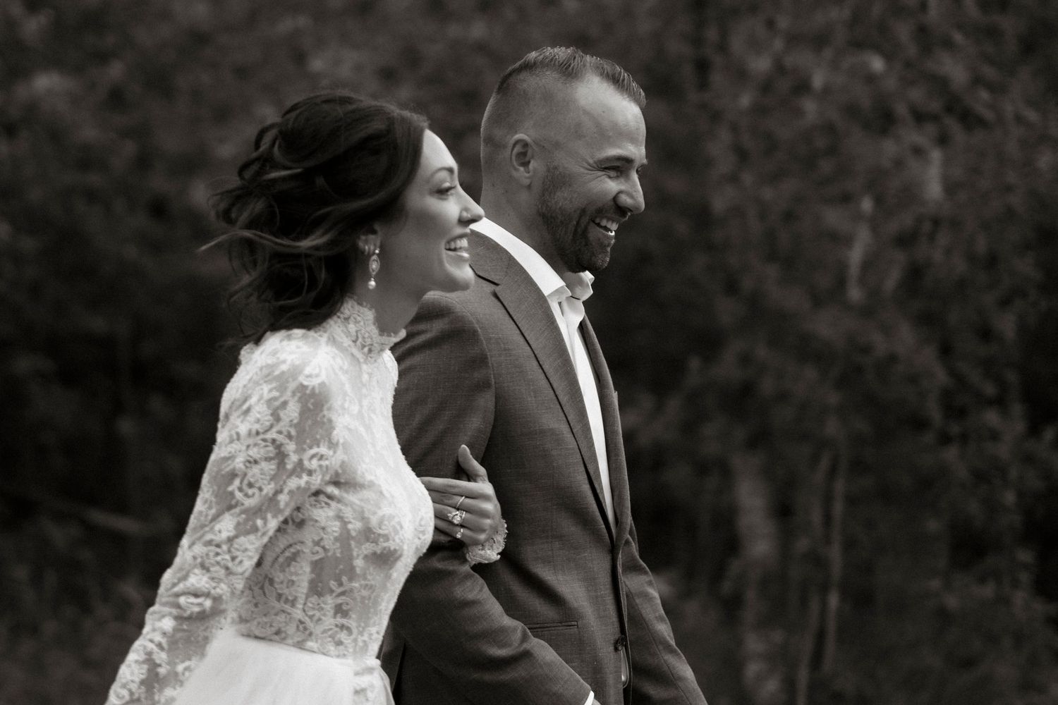 Black and white photo of a smiling bride in lace gown and groom in suit walking together outdoors.