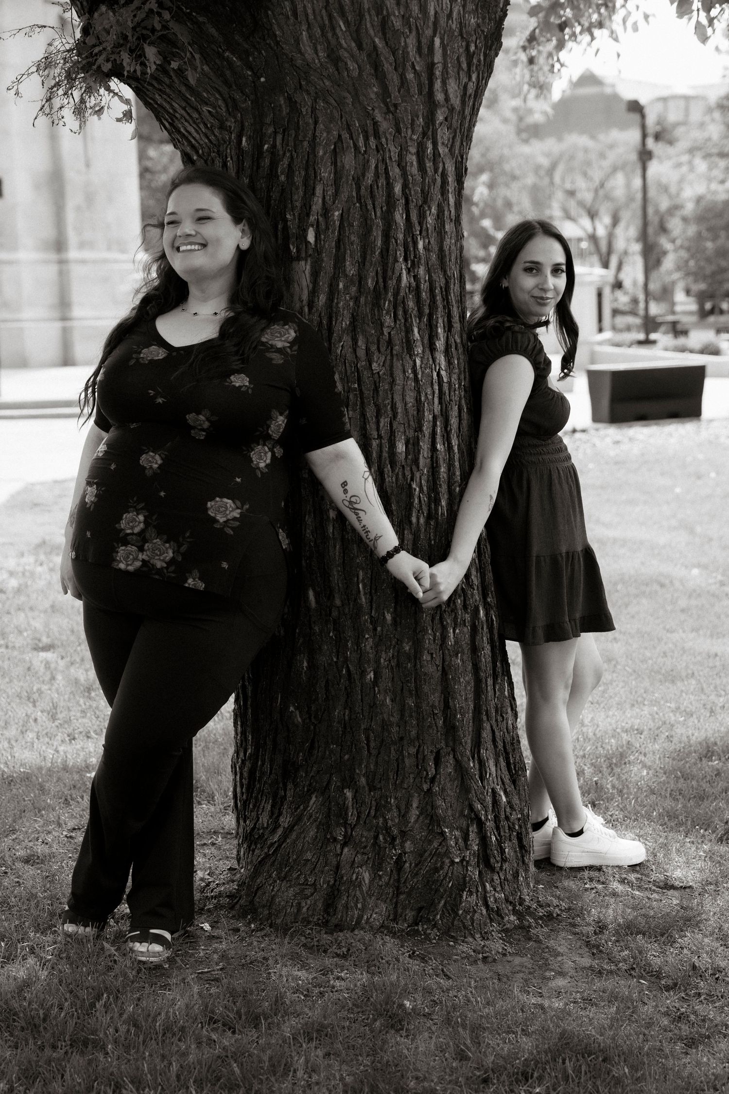Two women holding hands beside a large tree in a park, black and white photo.