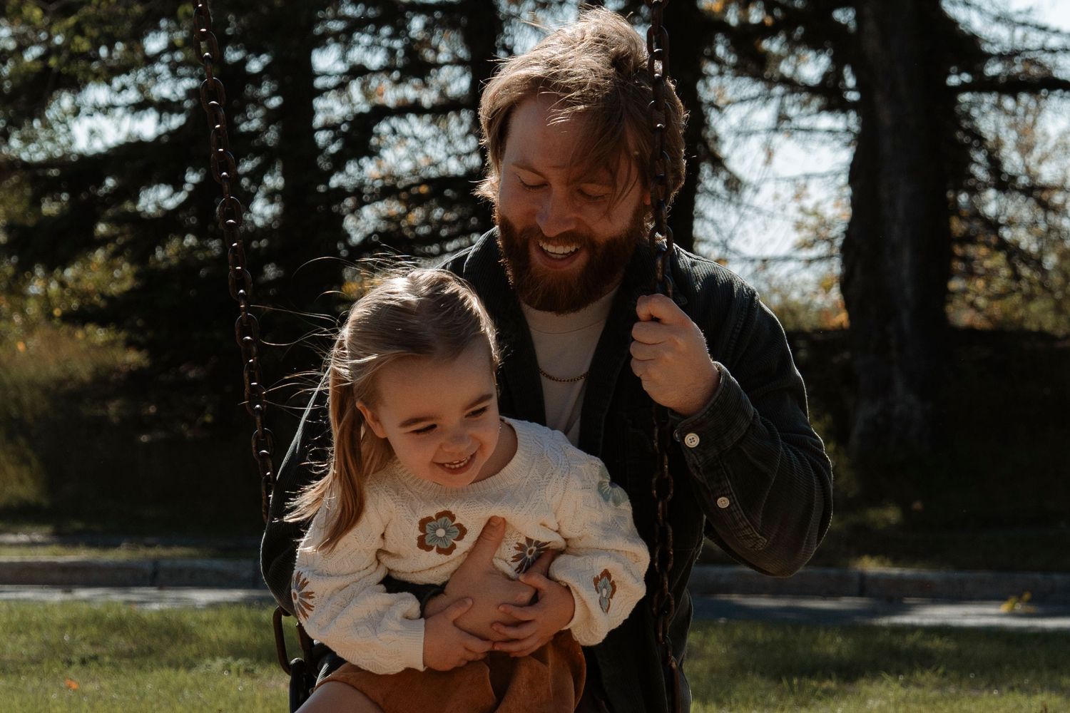A bearded father hugs his laughing young daughter on a swing in a sunny outdoor park setting.Family photography in Regina Saskatchewan.Famil