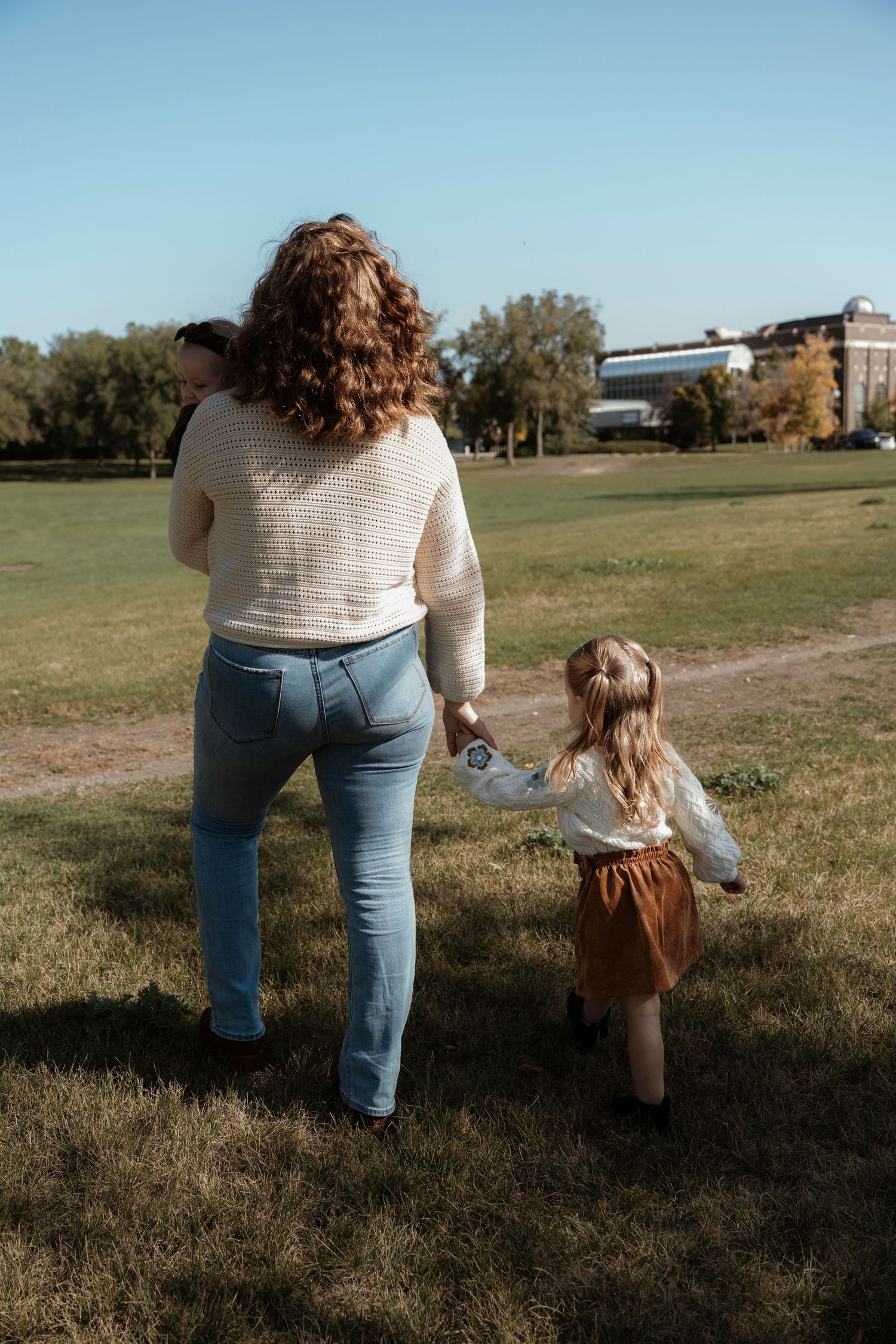 A mother with curly red hair walks hand-in-hand with her young daughter across a sunny green park while carrying a baby.Family photography.