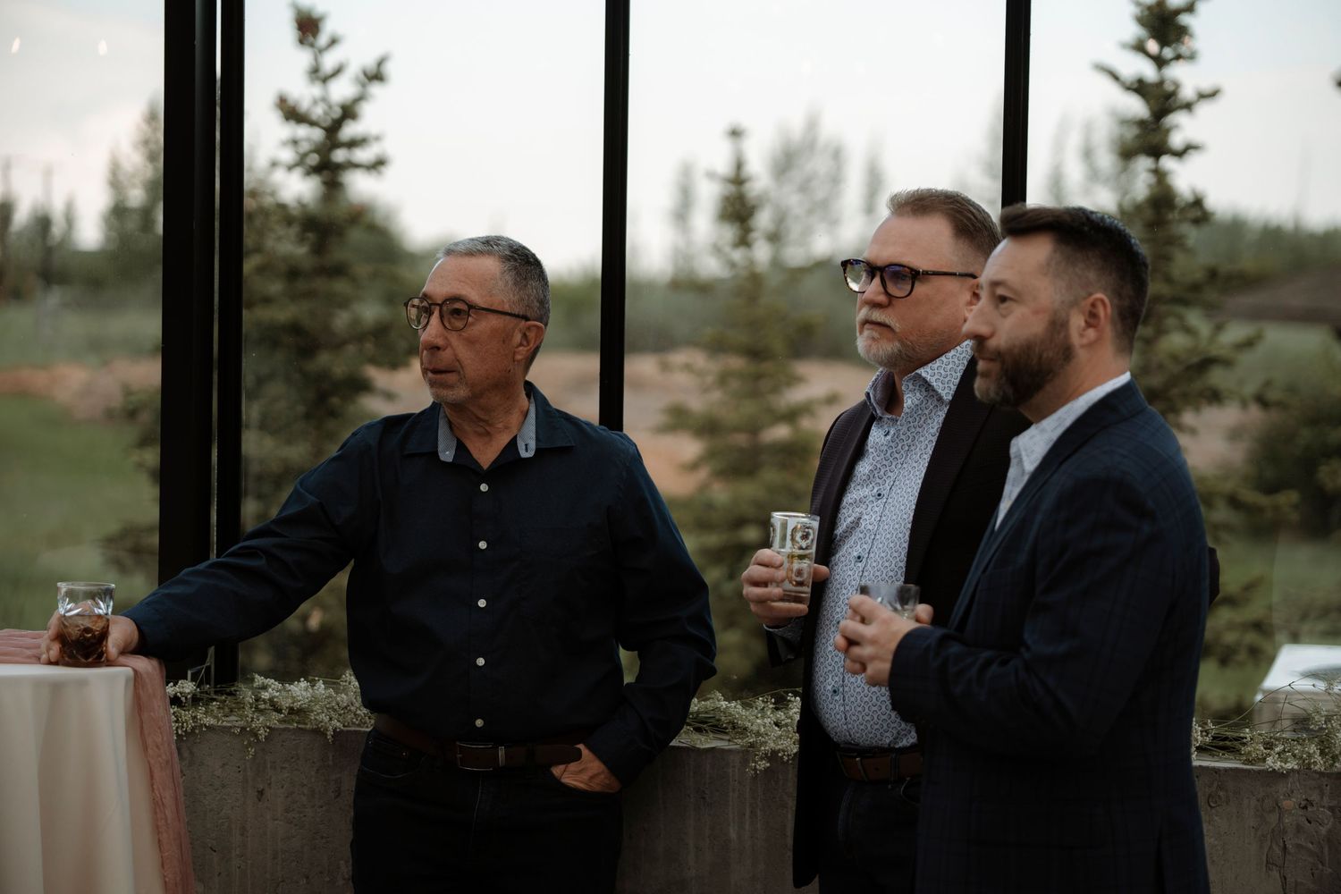 Three men in smart casual attire converse near large windows overlooking a forested landscape at an evening event.