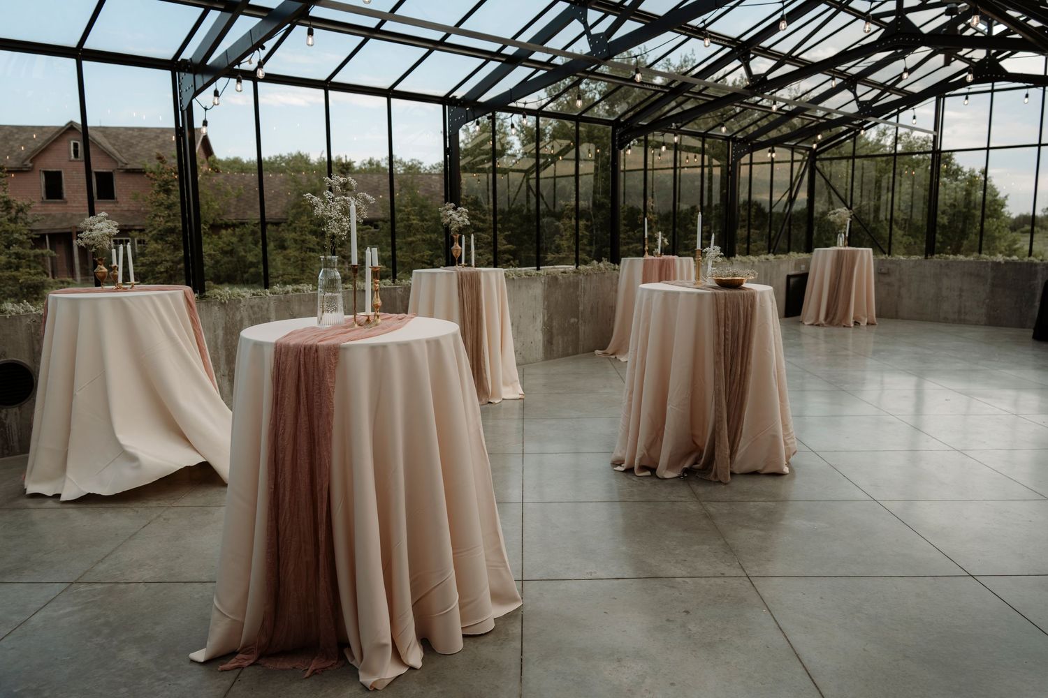 Elegant greenhouse venue with blush pink cocktail tables set for a wedding reception, surrounded by lush greenery.