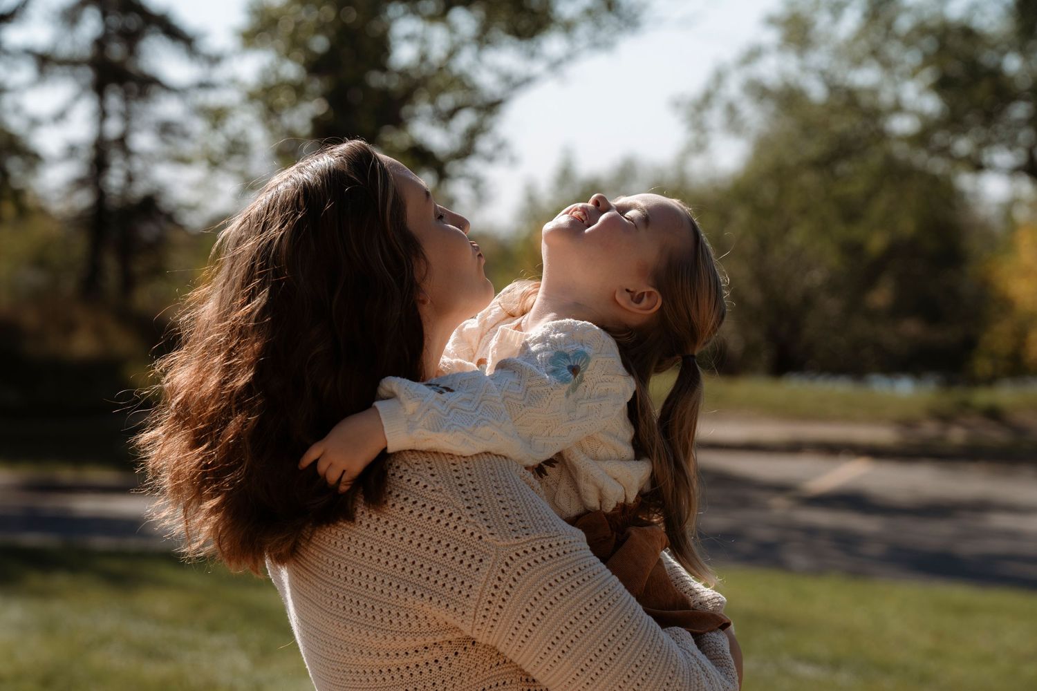 A mother in a cream sweater joyfully lifts her smiling young daughter in a sunny outdoor park.Family photography in Regina Saskatchewan.