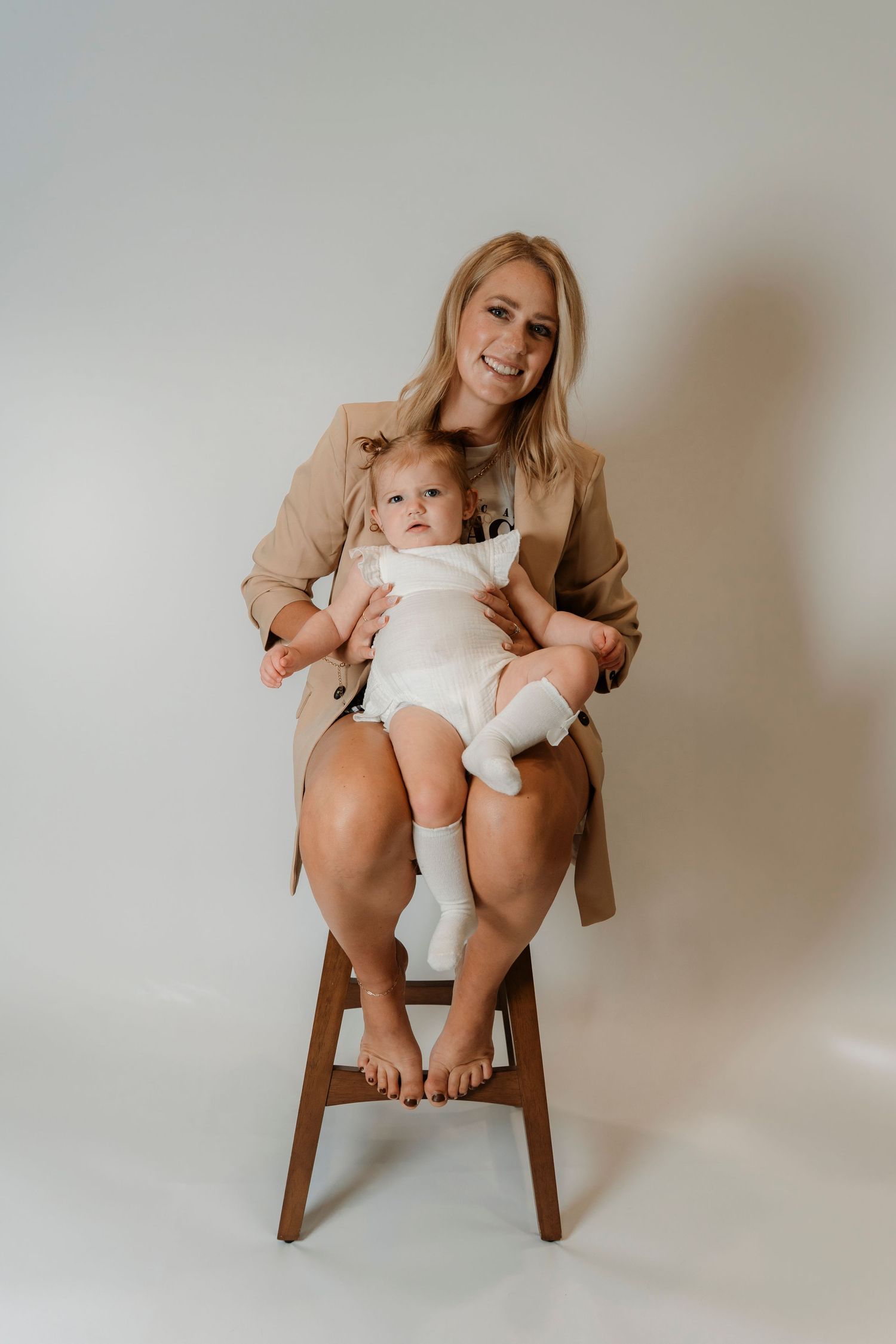 Smiling blonde woman in beige blazer sitting on wooden stool holding a baby in white onesie against light background.