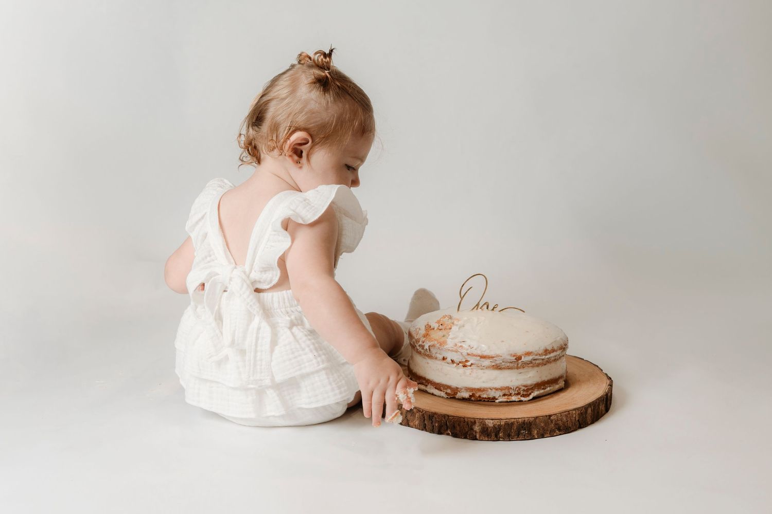 Baby girl in white ruffled dress reaching toward a naked smash cake on a wooden slice.Family photography in Regina Saskatchewan.