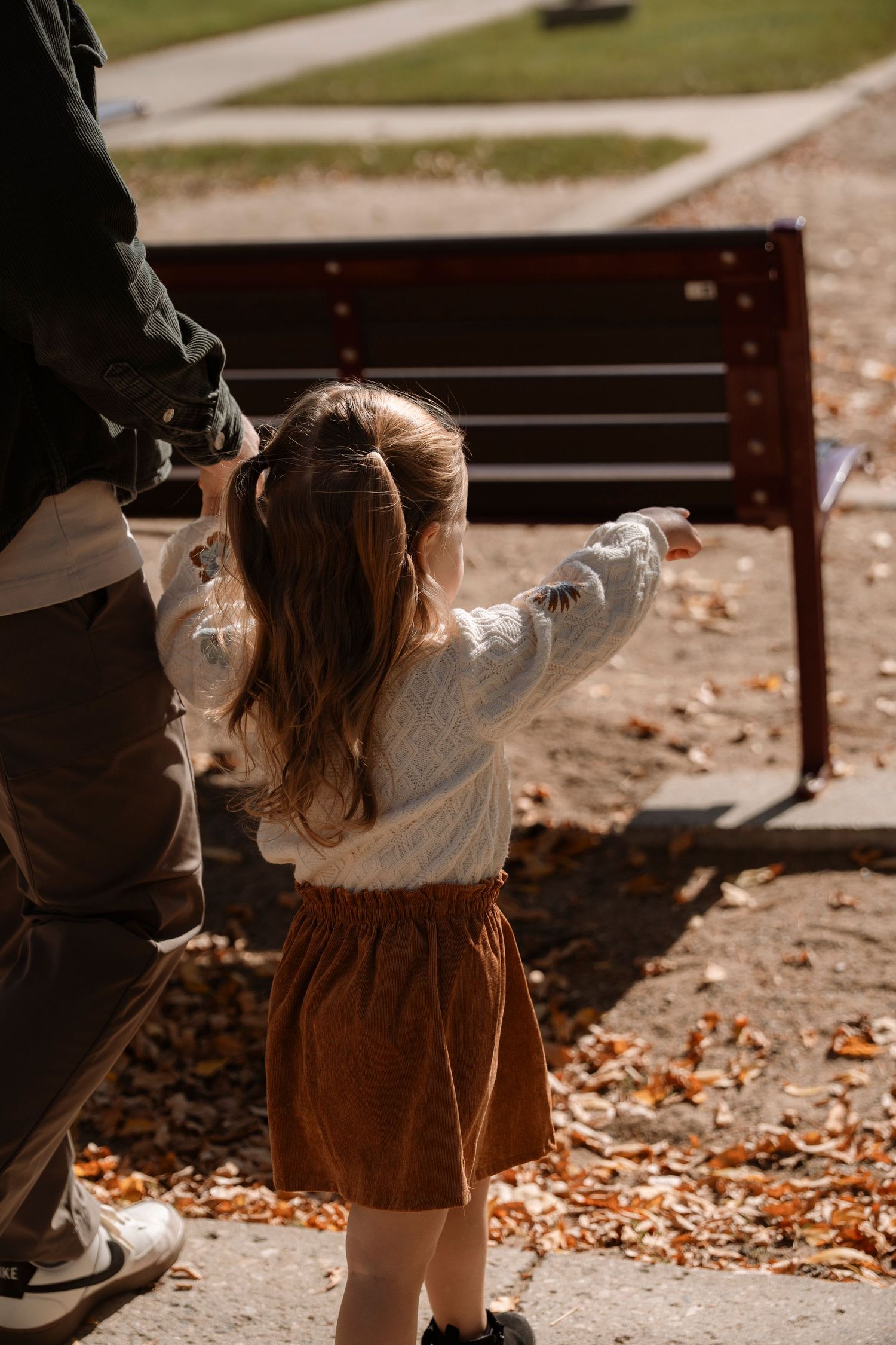 Young girl in white sweater and brown skirt reaching toward a park bench while holding an adults hand.Family photography in Regina Sask.