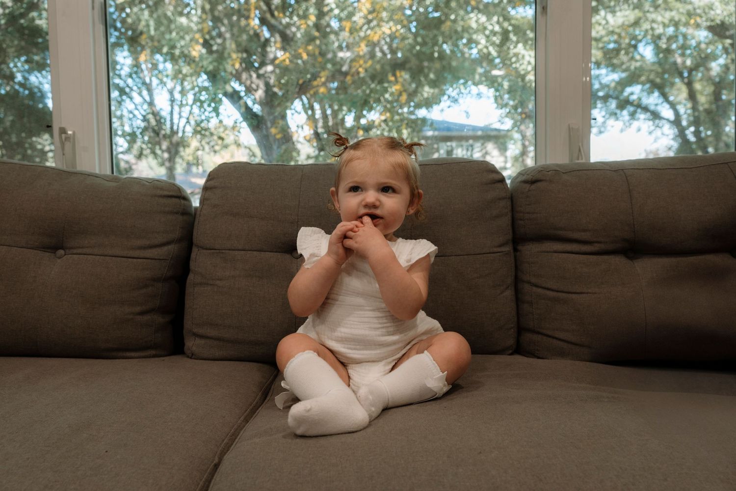 Cute baby girl in white outfit sitting on a gray couch near a large window.Family photography in Regina Saskatchewan.