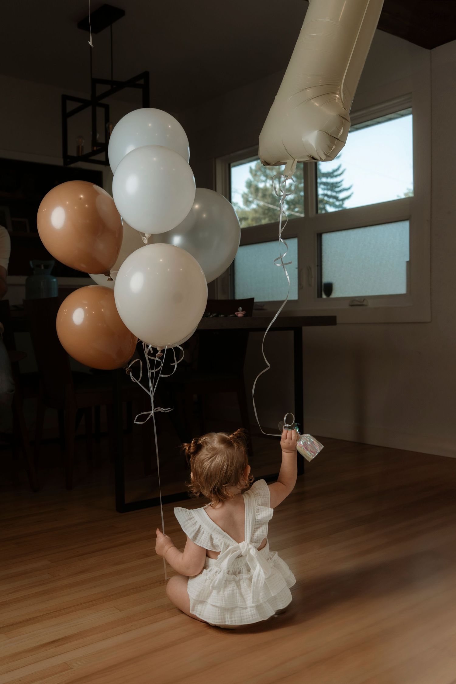 A baby in a white dress sits on hardwood floor holding brown, white, and gold birthday balloons.Family photography in Regina Saskatchewan.