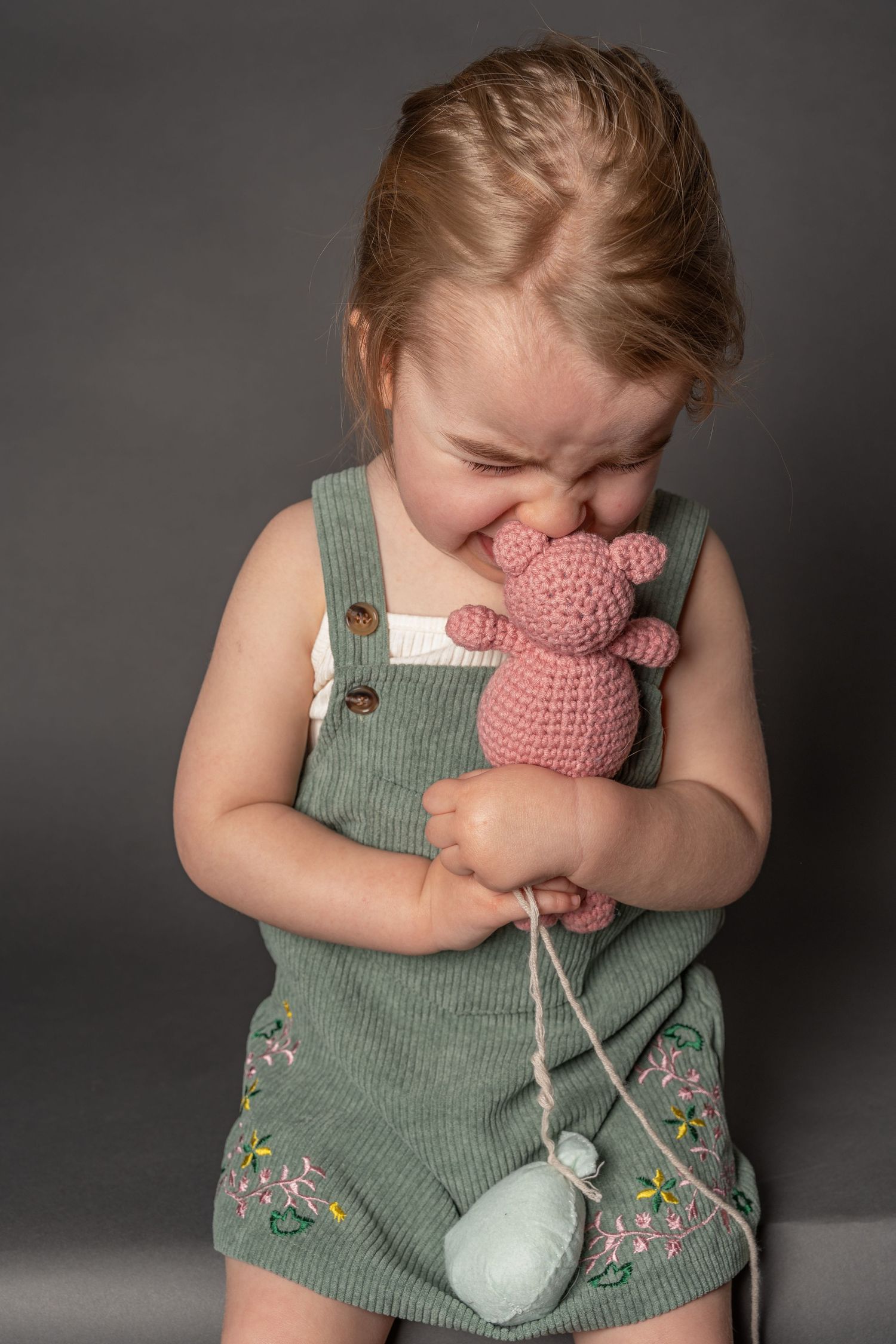 Toddler girl in green overalls hugging a pink crocheted teddy bear toy, smiling with eyes closed.