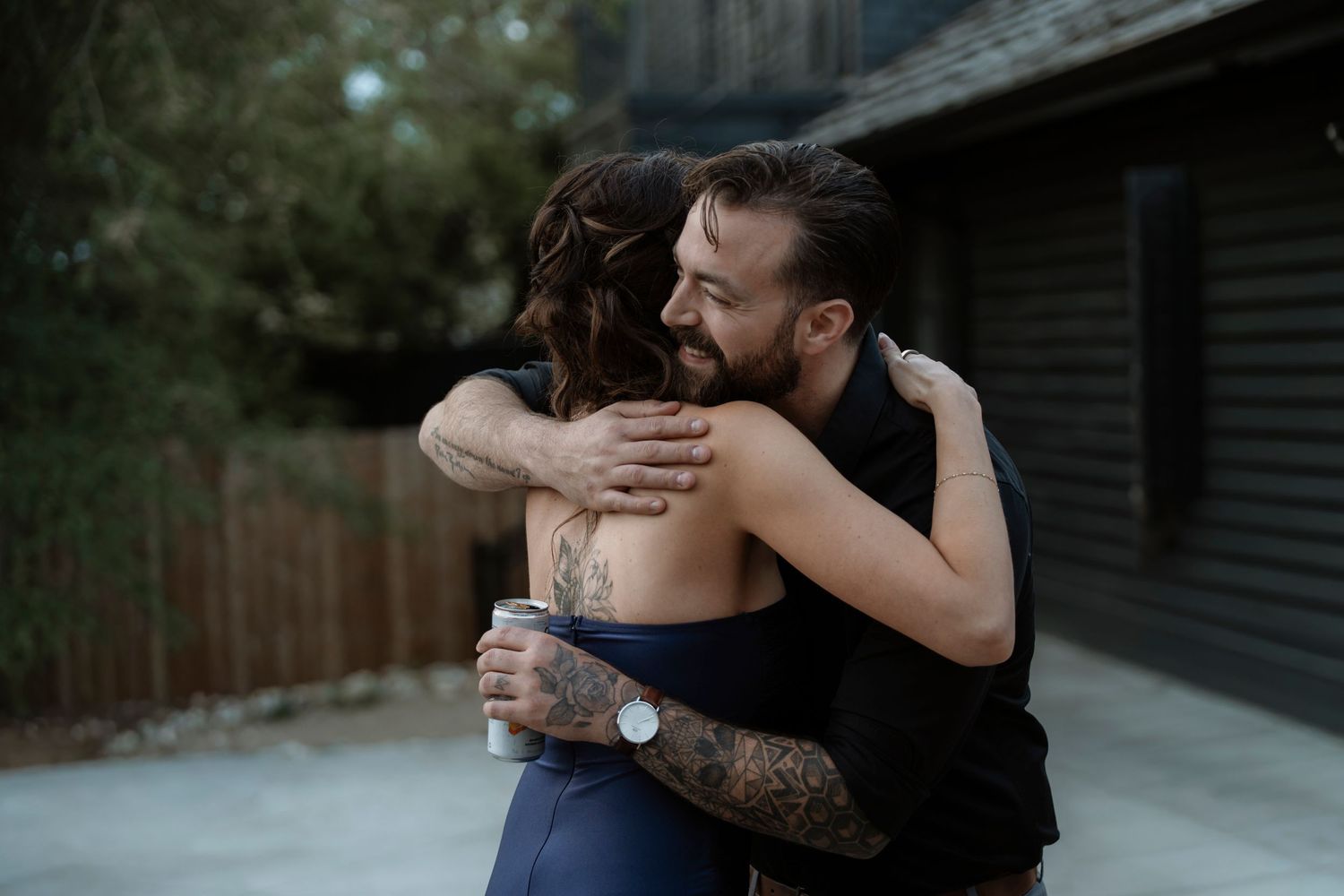 A smiling tattooed man hugs a woman with a back tattoo outdoors near a wooden fence and dark building.