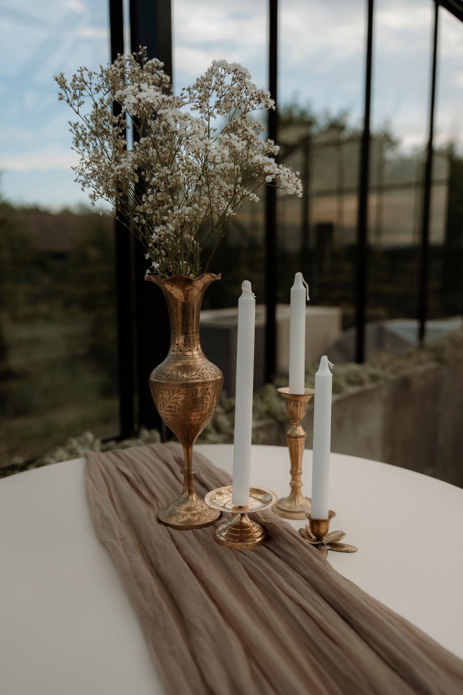 Gold brass vase with baby's breath and white taper candles on candlesticks atop a white table with beige runner.