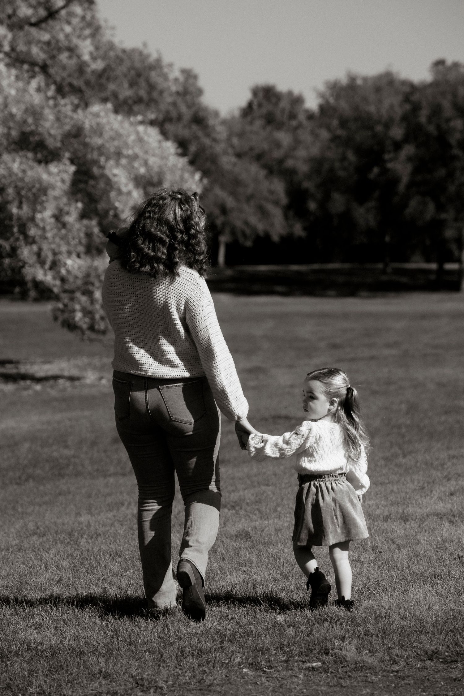 A mother and young daughter walk hand-in-hand across a grassy park in this classic black and white photograph. Family photography in Regina