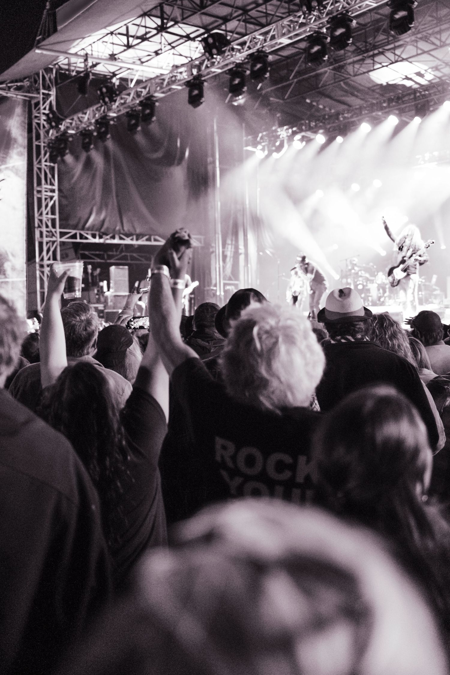 Black and white photo of a rock concert crowd with hands raised, stage lights blazing, fan wearing Rock You shirt visible.