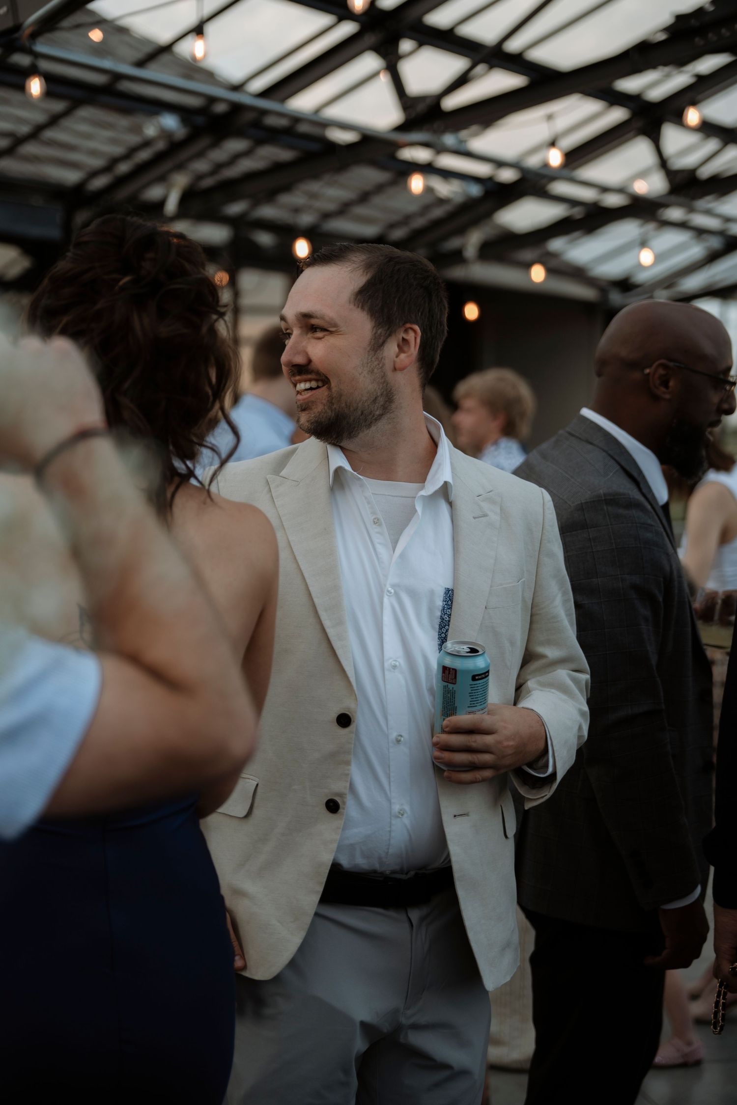 A smiling man in a beige blazer holds a drink can at an outdoor wedding reception with string lights.
