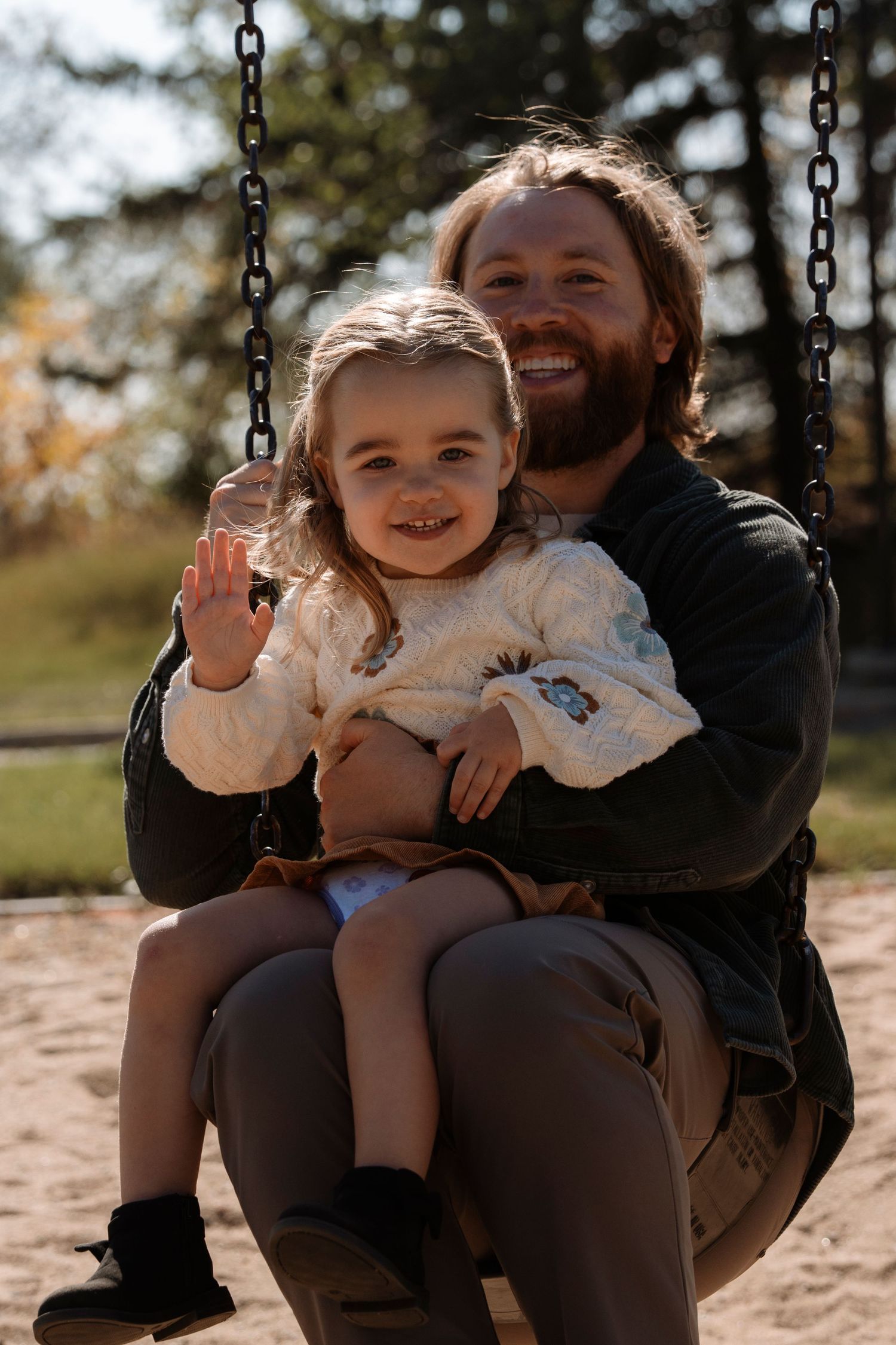 A smiling bearded father sits on a swing holding his happy young daughter at an outdoor playground. Family photography in Regina Sask.