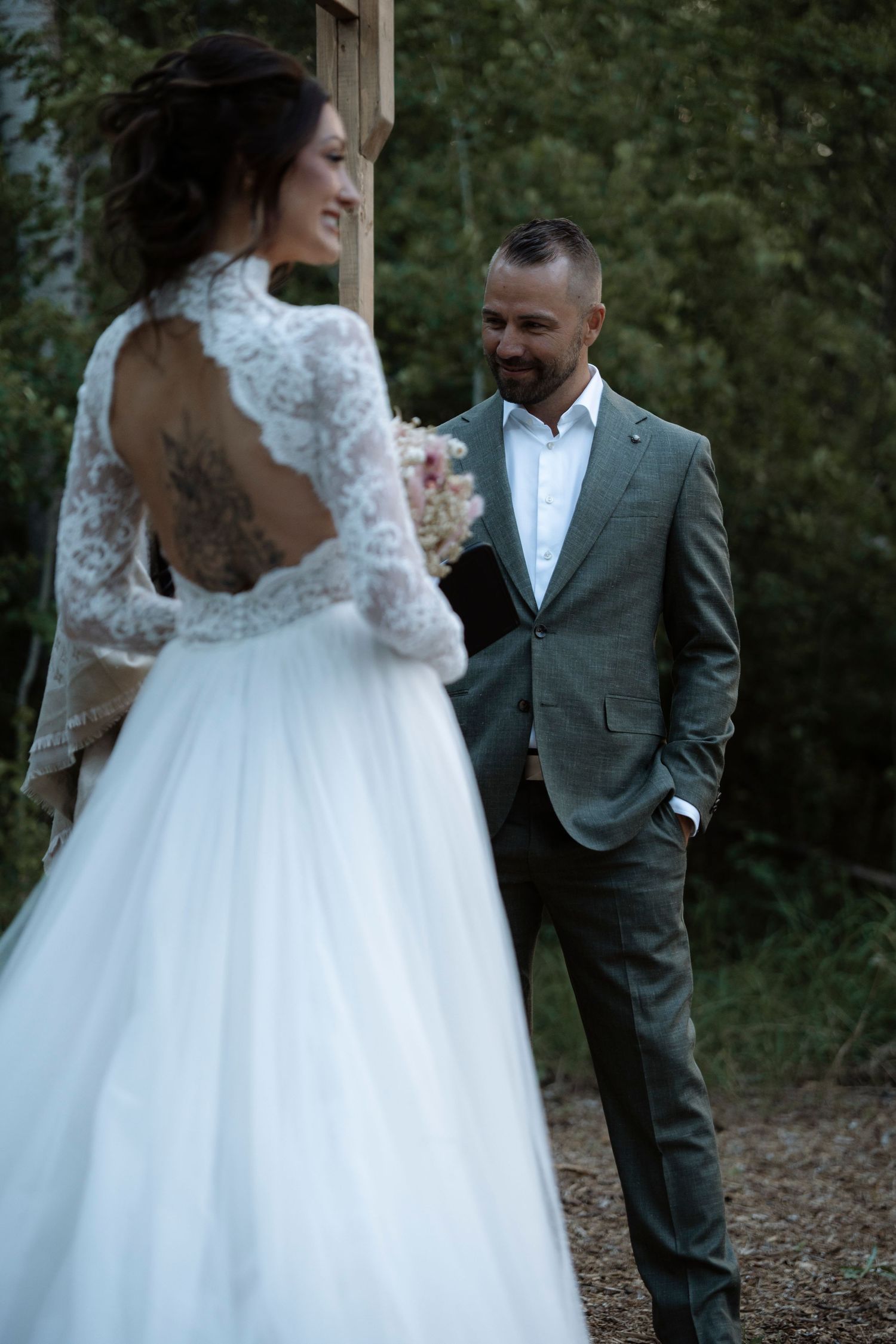 Groom in gray suit smiles at bride in lace backless wedding dress with tattoo, holding bouquet outdoors.