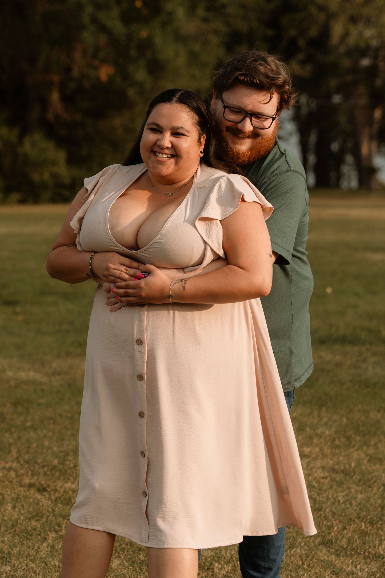 A smiling couple embraces outdoors in a park, woman in a cream button-down dress, man in a green shirt.