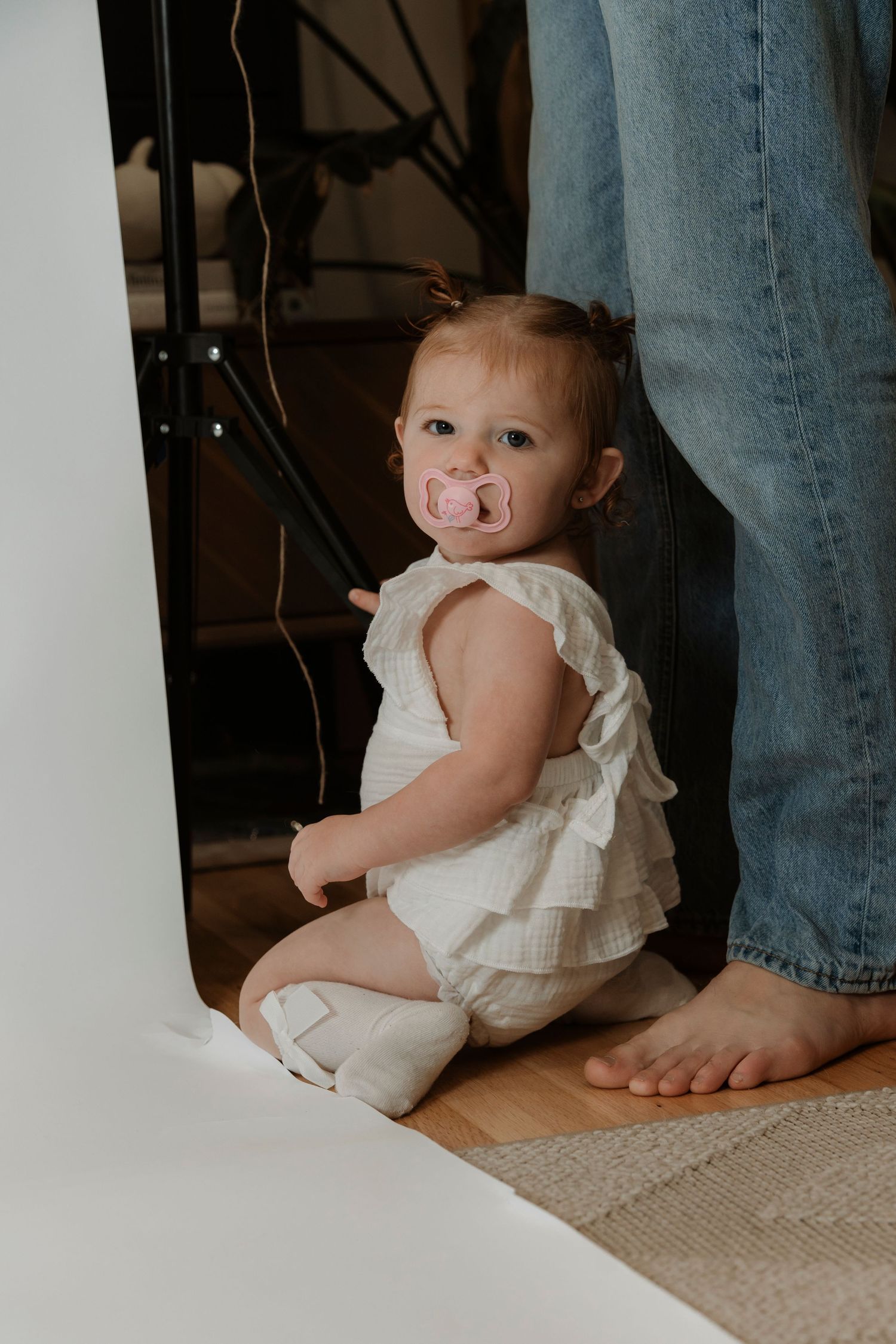 Baby girl with pink pacifier sitting on floor beside a photography backdrop and adult feet.Family photography in Regina Saskatchewan.