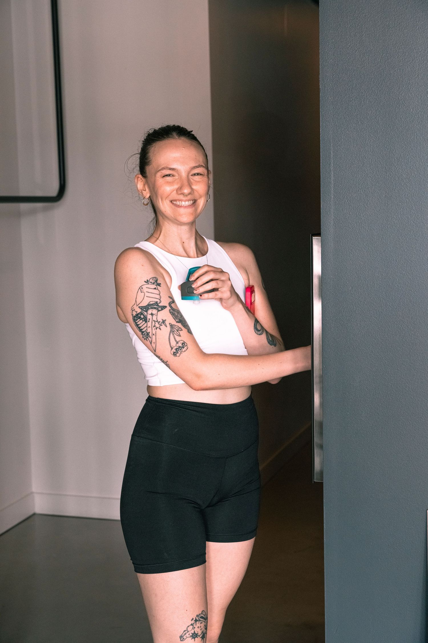 Smiling tattooed woman in white crop top and black shorts holding a phone in a modern gym.
