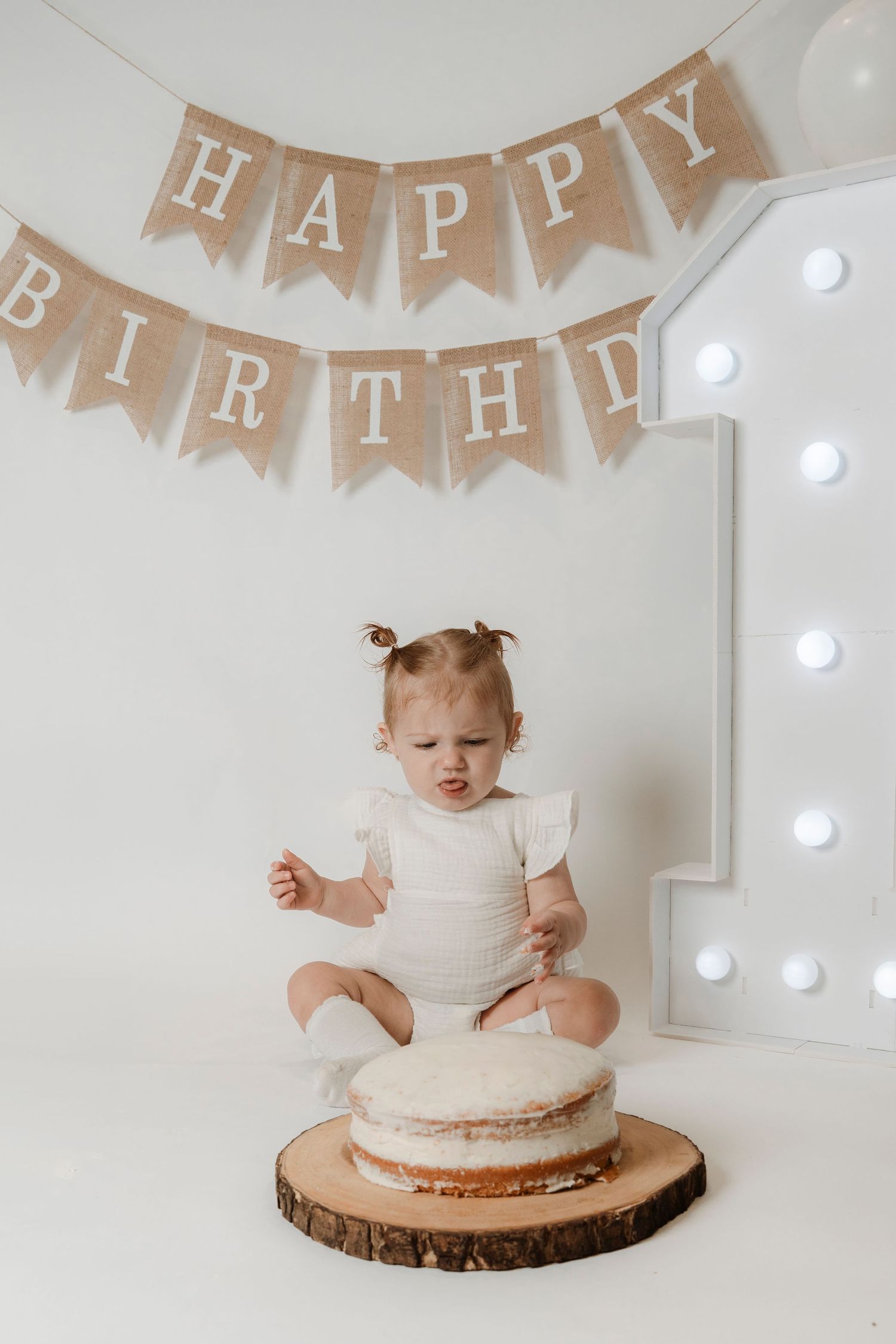 A baby girl smashes her first birthday cake under a burlap Happy Birthday banner and number one.Family photography in Regina Saskatchewan.
