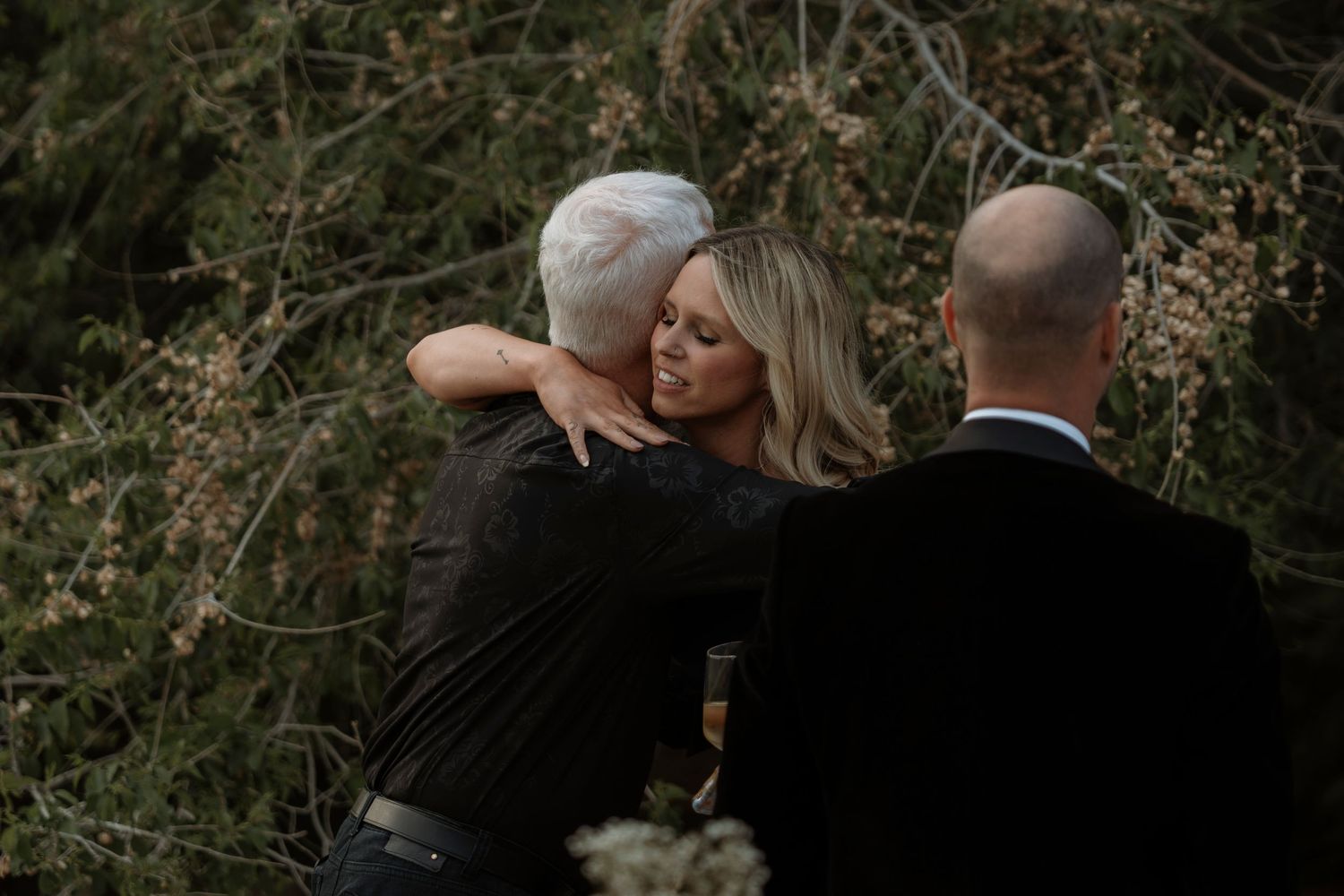 A blonde woman in black embraces an older man with white hair outdoors, while a bald man stands nearby among trees.