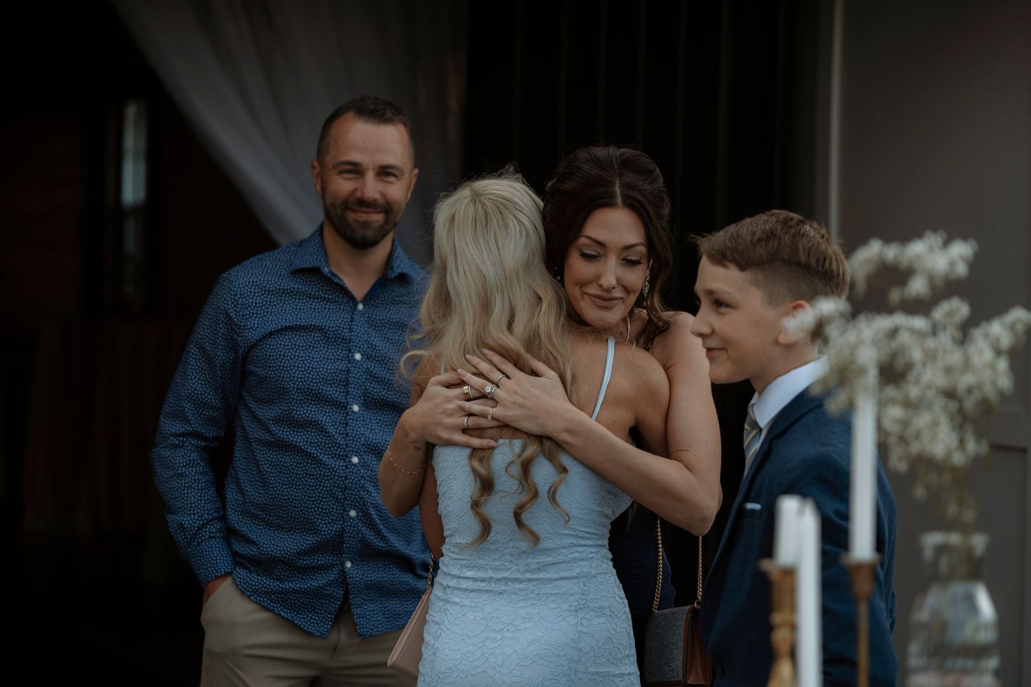 Two women embrace warmly at a wedding ceremony while a smiling man and young boy look on nearby.