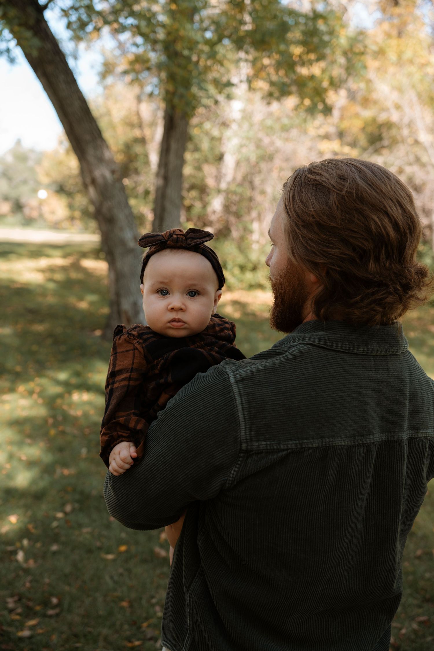 A bearded father holds his infant daughter in a brown plaid dress outdoors in a fall park setting.Family photography in Regina Saskatchewan.