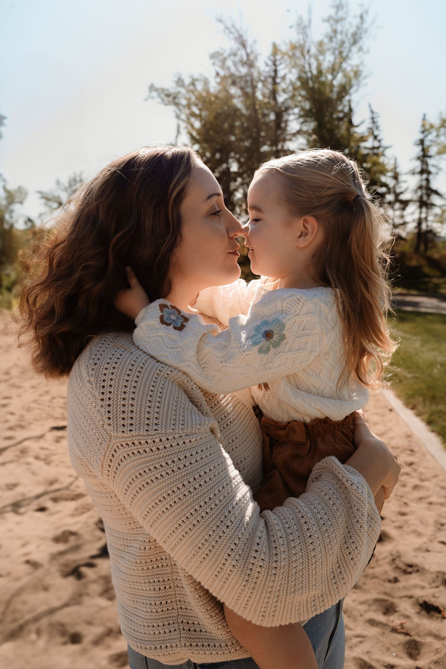 Mother and daughter sharing a tender nose-to-nose moment outdoors on a sandy beach in matching cream knit sweaters.Family photography.