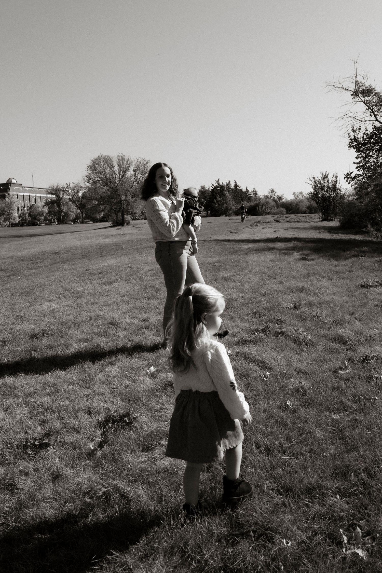 A mother lifts her baby while her young daughter stands beside her in a park, captured in black and white. Family photography in Regina.
