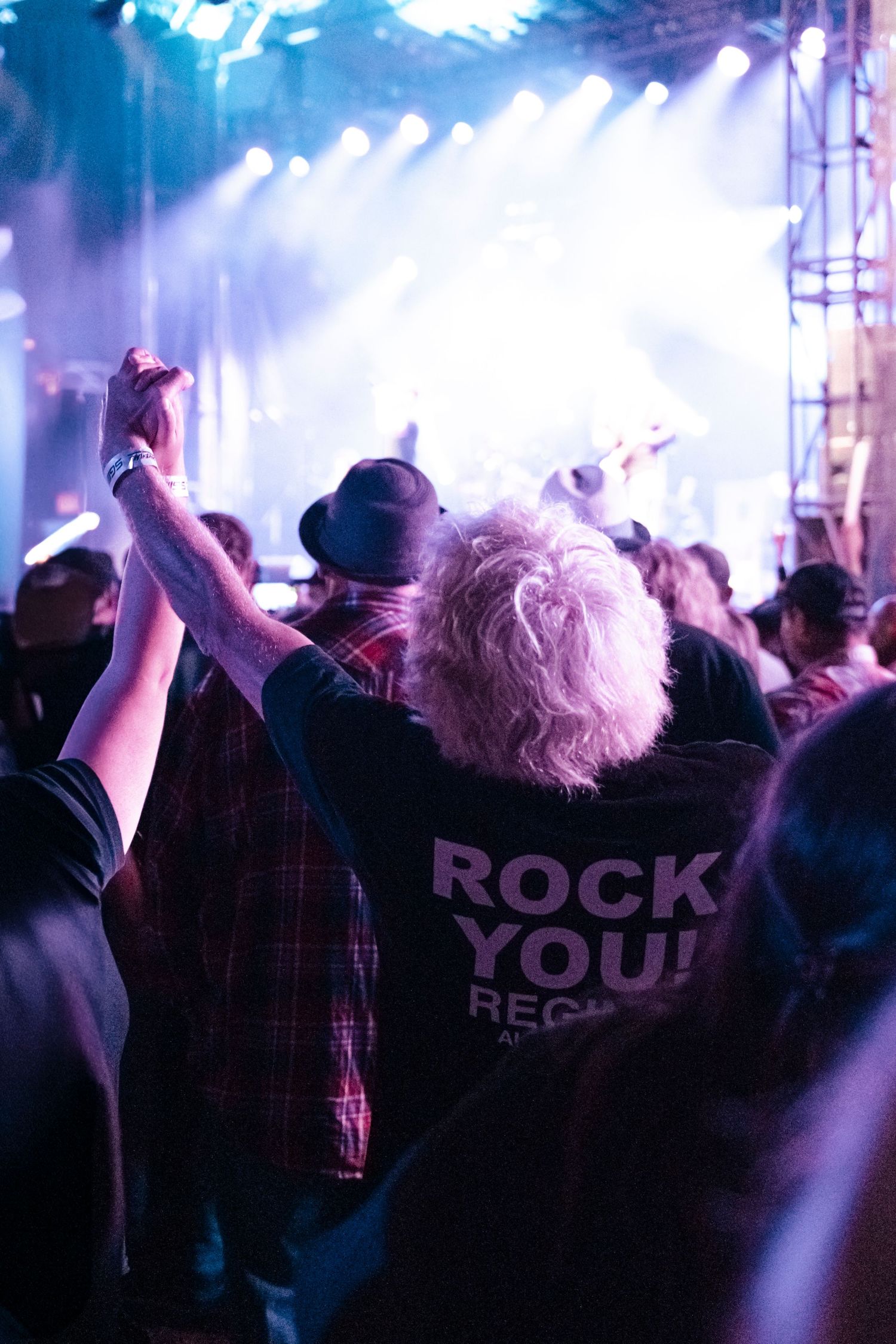 Crowd at a rock concert, person in black Rock You t-shirt with white hair raises fist toward bright stage lights.