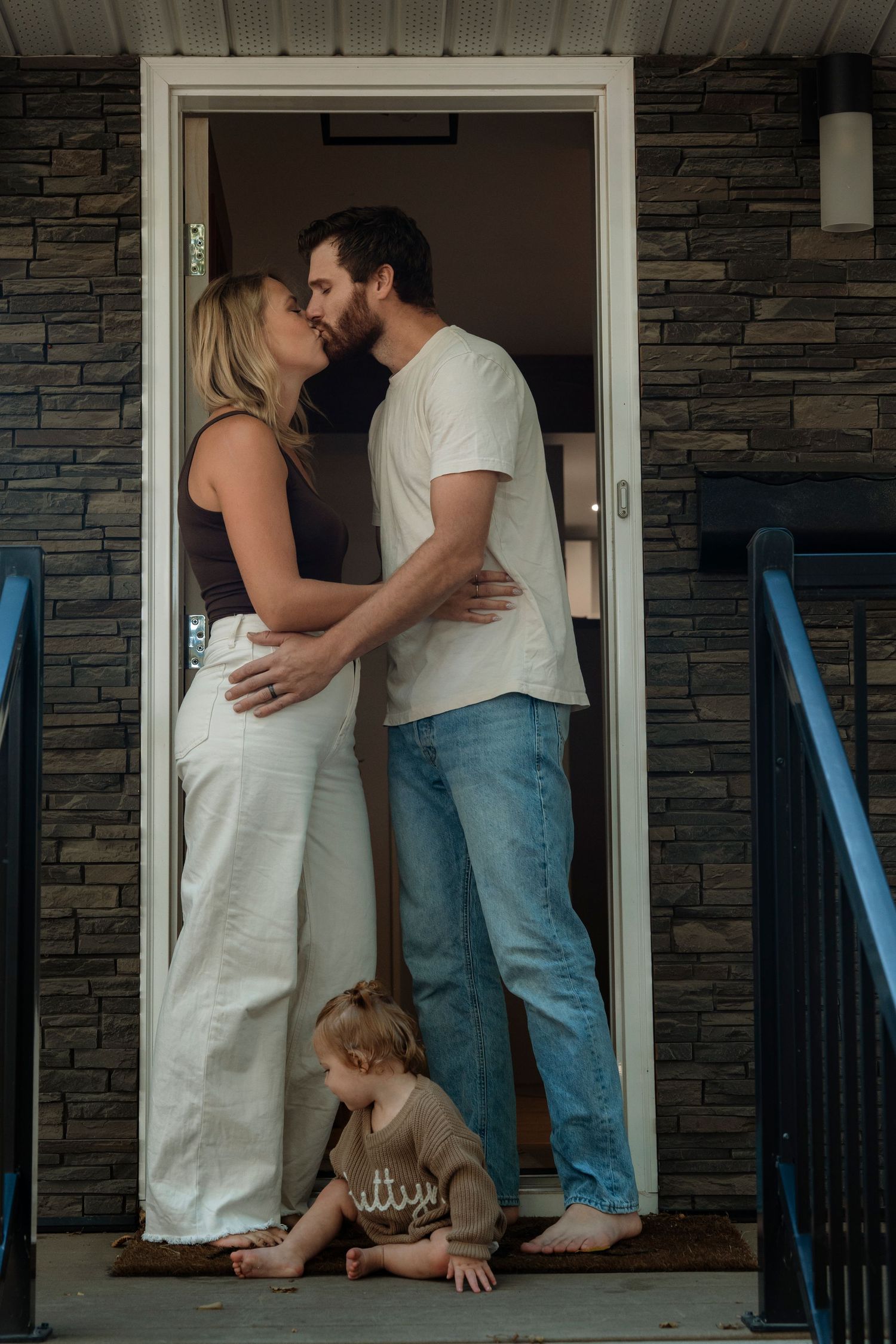 Couple sharing a kiss in doorway while their toddler sits on the front porch below.Family photography in Regina Saskatchewan.
