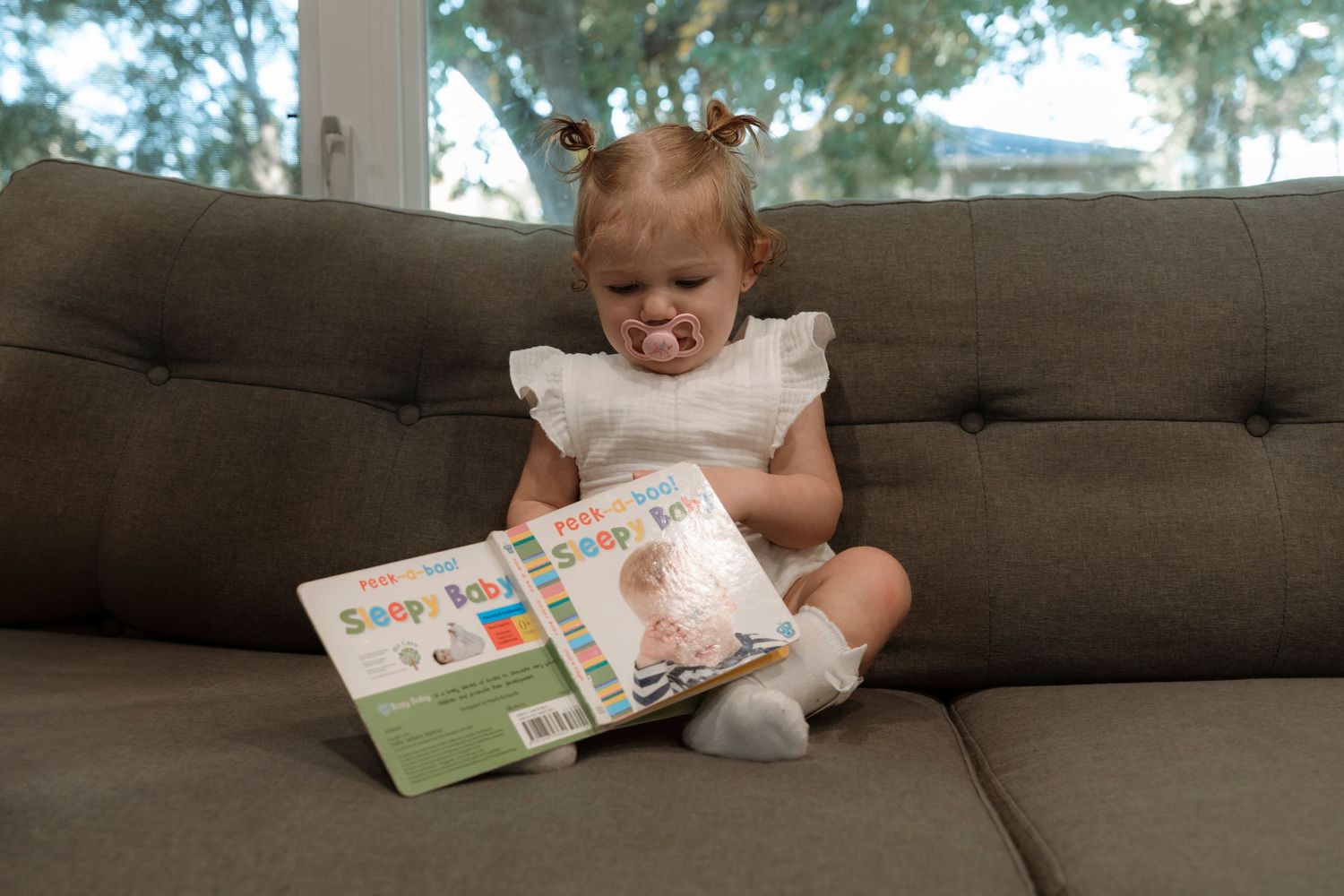 Cute toddler girl sitting on gray couch reading a Sassy Baby board book. Family photography in Regina Saskatchewan.