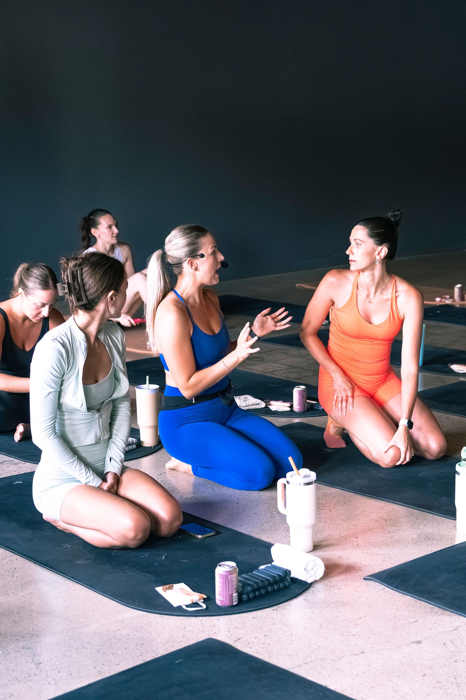 Women in activewear sitting on yoga mats chatting after a fitness class in a modern studio.