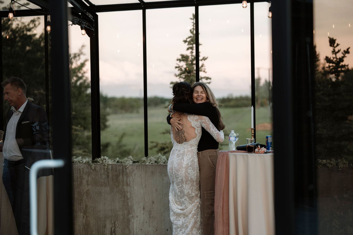Bride in lace wedding gown shares a warm embrace with a guest at an elegant outdoor wedding reception venue.