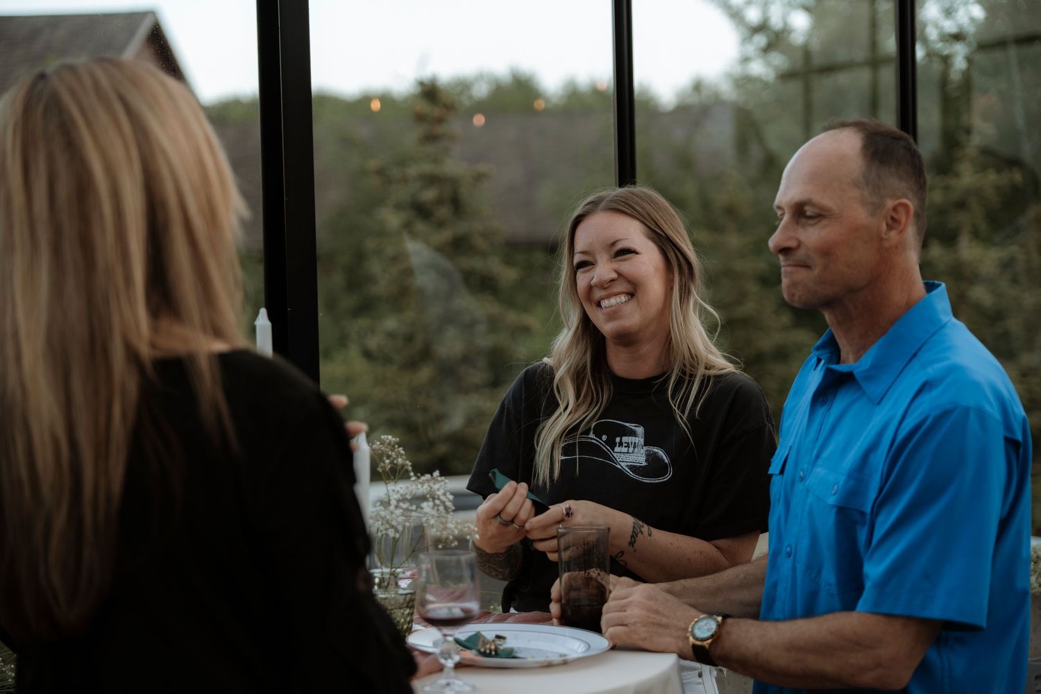 Three people smiling and laughing together at a dining table in a glass-enclosed room during an evening gathering.