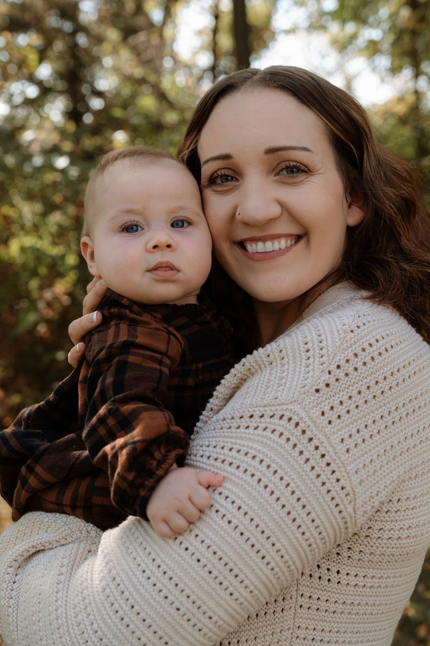 Smiling brunette mother in white knit sweater holding her baby boy in autumn forest.Family photography in Regina Saskatchewan.