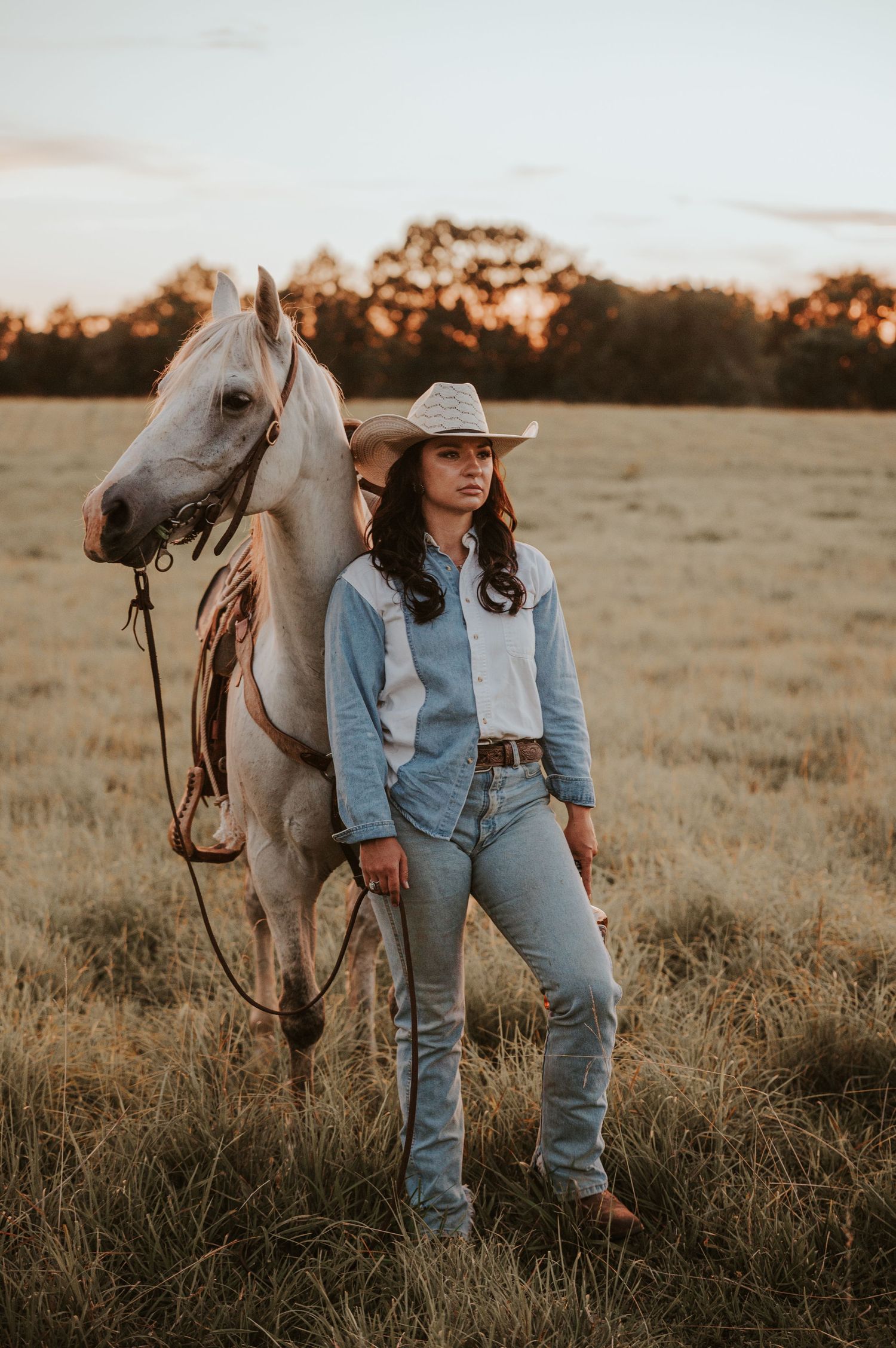 Denim-clad figure standing beside a white horse in a golden sunset field.