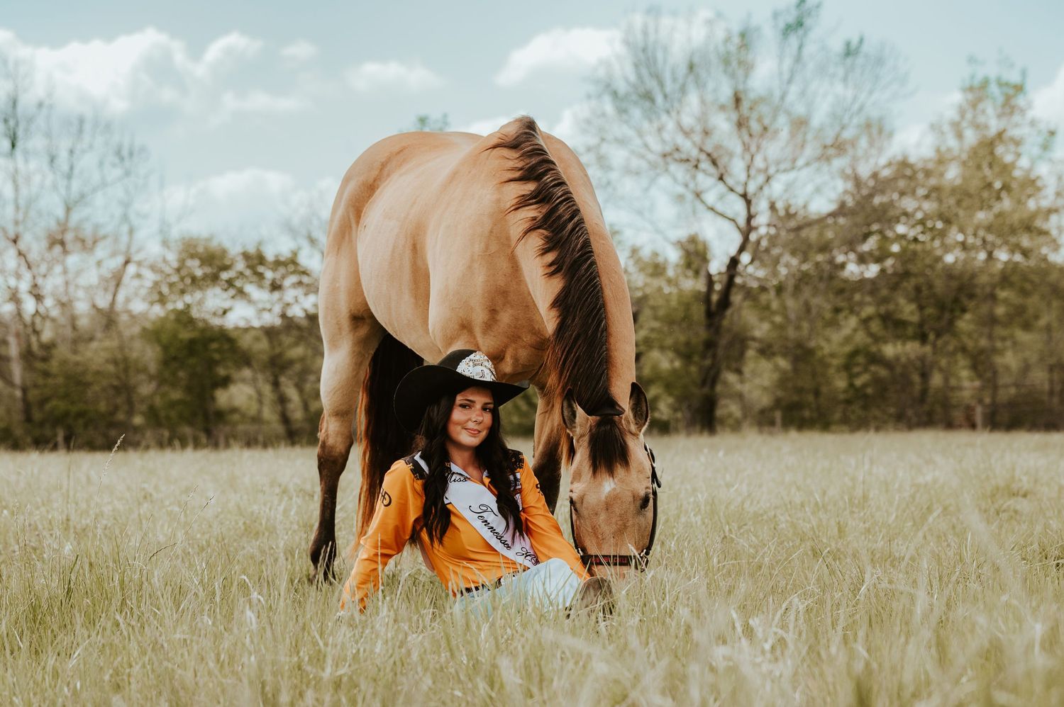 A smiling woman in a cowboy hat sits in a grassy field beside a grazing buckskin horse under a cloudy sky.