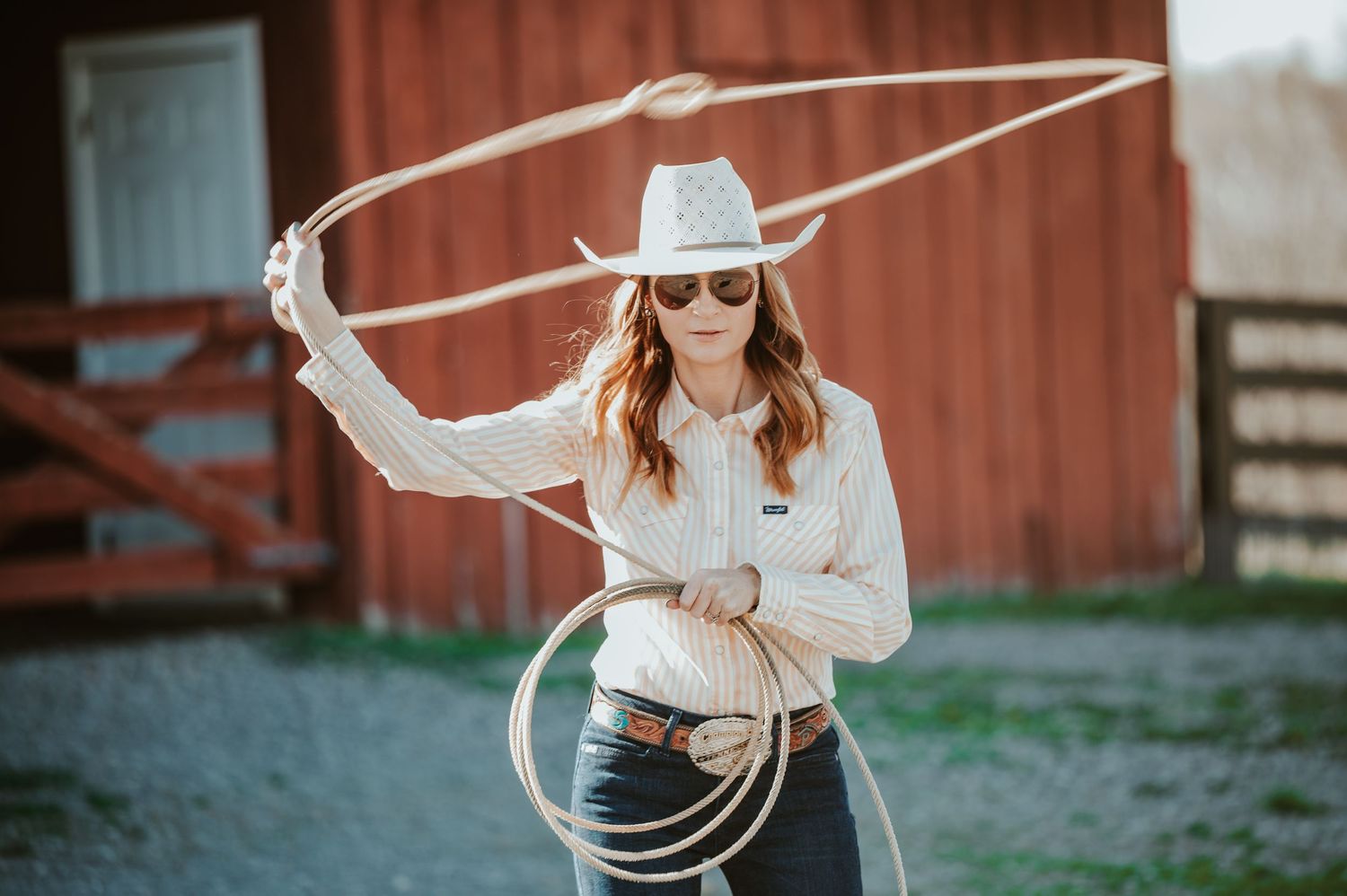 Person in western wear demonstrating rope tricks against red barn backdrop.