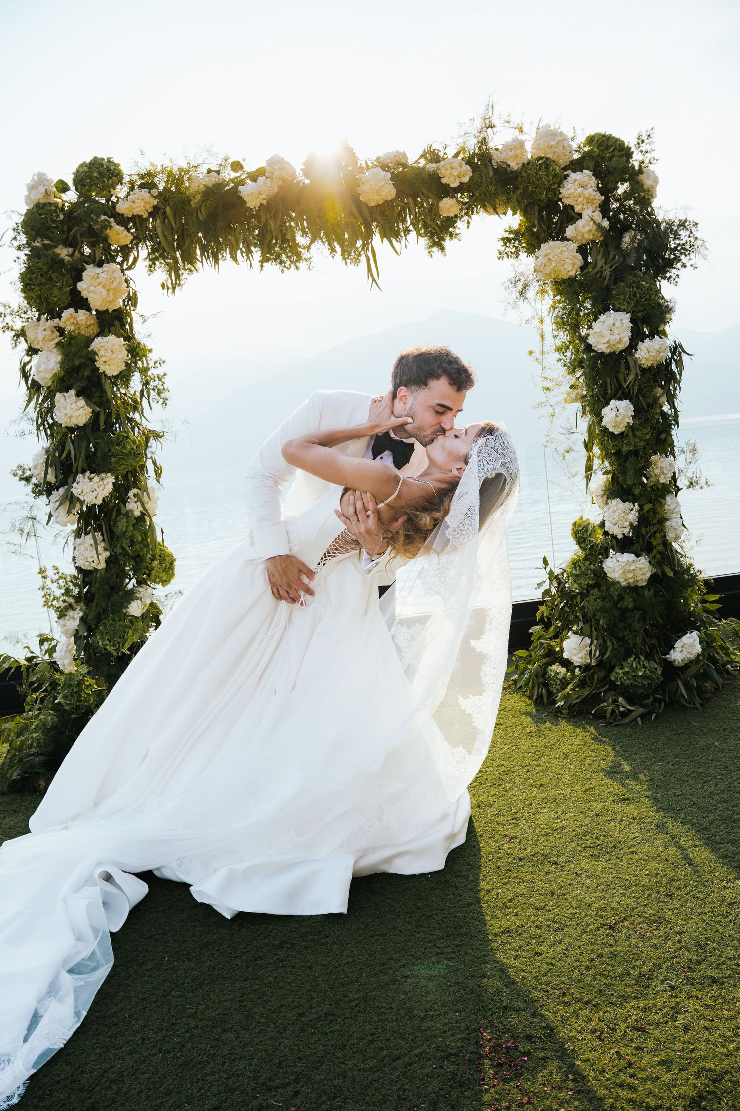 Bride and groom share a romantic dip kiss under a floral arch adorned with white roses and greenery at an outdoor wedding.