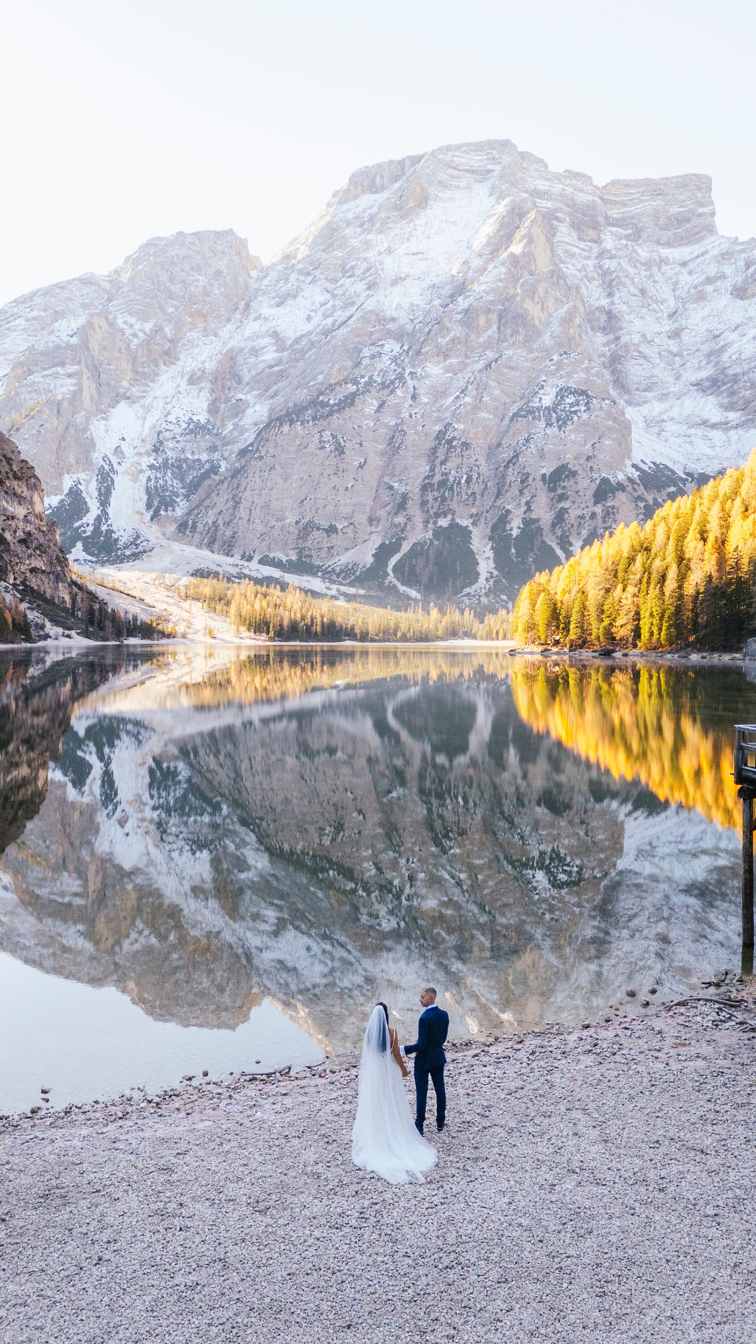 Two people admire the stunning reflections of snow-capped mountains in Lago di Braies during golden hour in the Dolomites.