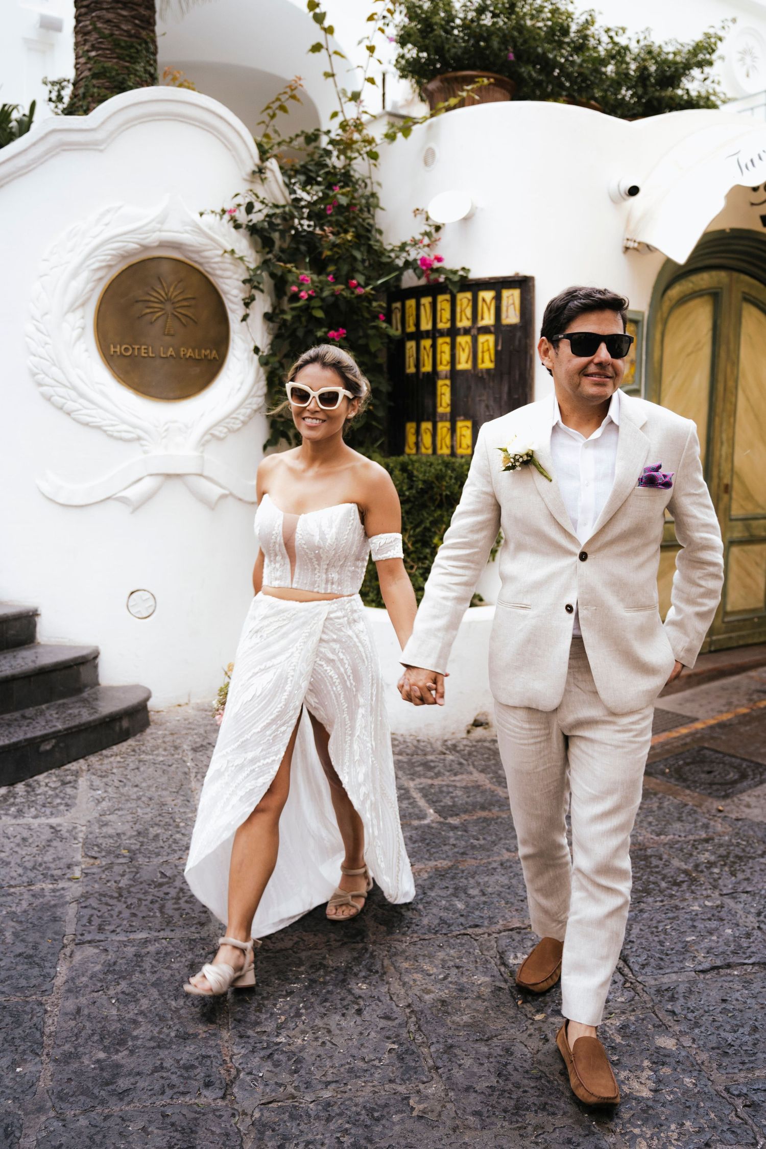 A couple in all-white wedding attire walks together on a cobblestone street holding hands.