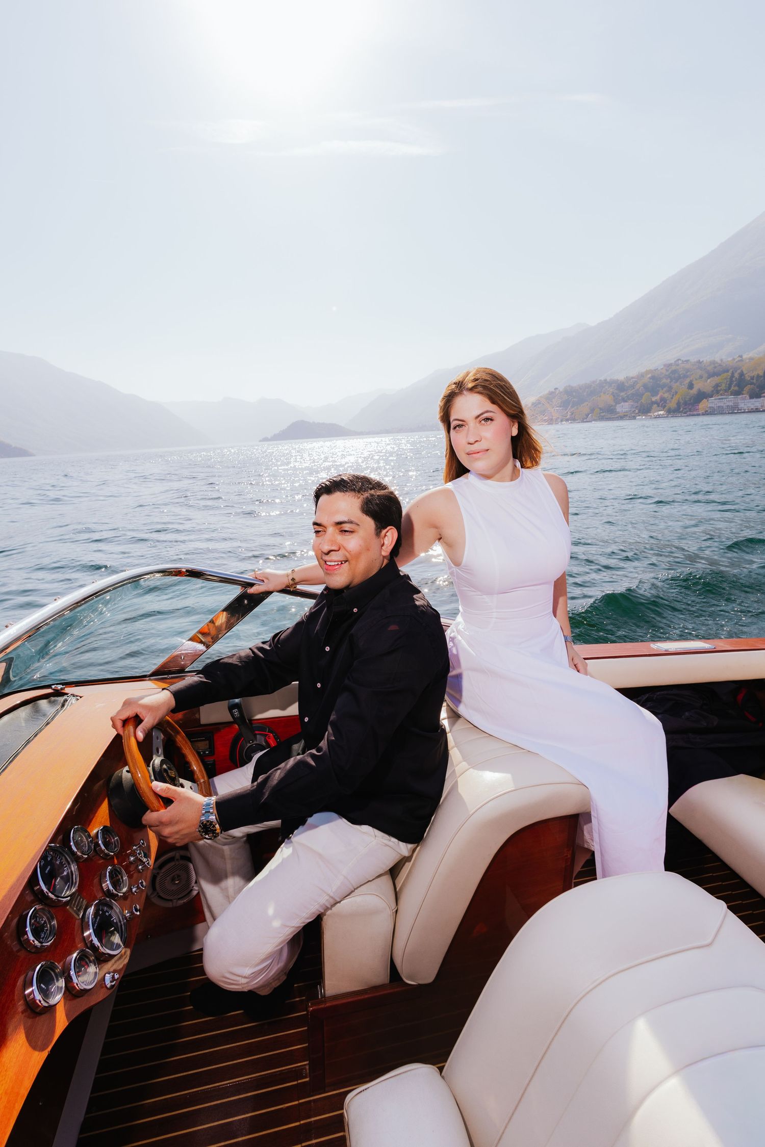 Couple in wedding attire on a luxury wooden speedboat on a scenic mountain lake, bride in white dress, groom in black suit.