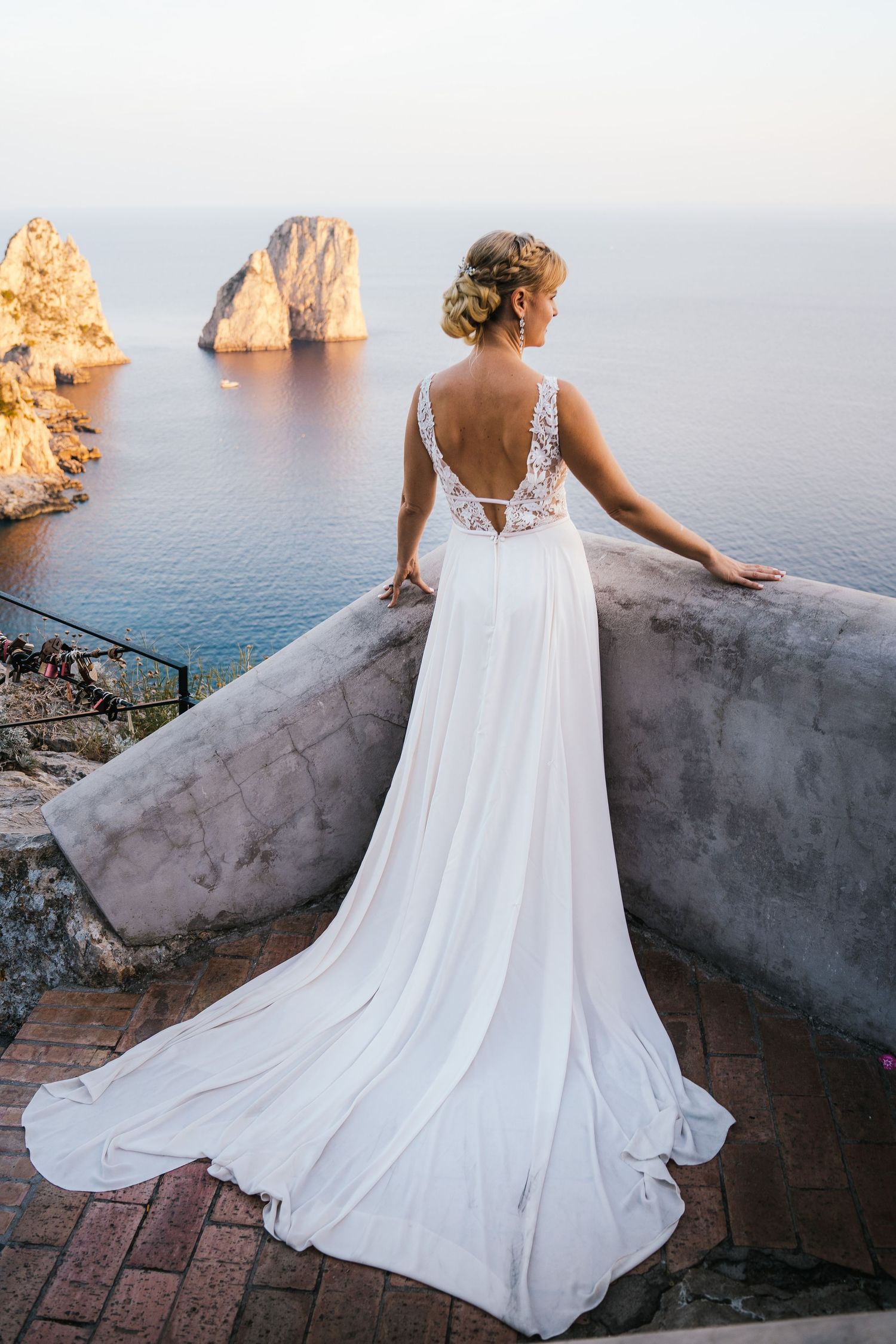 A bride in a flowing white wedding dress stands at a scenic overlook with Mediterranean sea cliffs and rock formations at sunset.