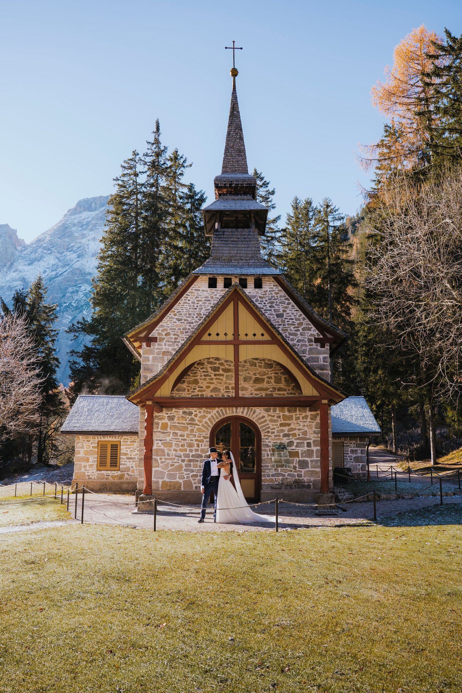 Small rustic wooden chapel nestled in a mountain meadow with snow-capped peaks and evergreen trees in the background.